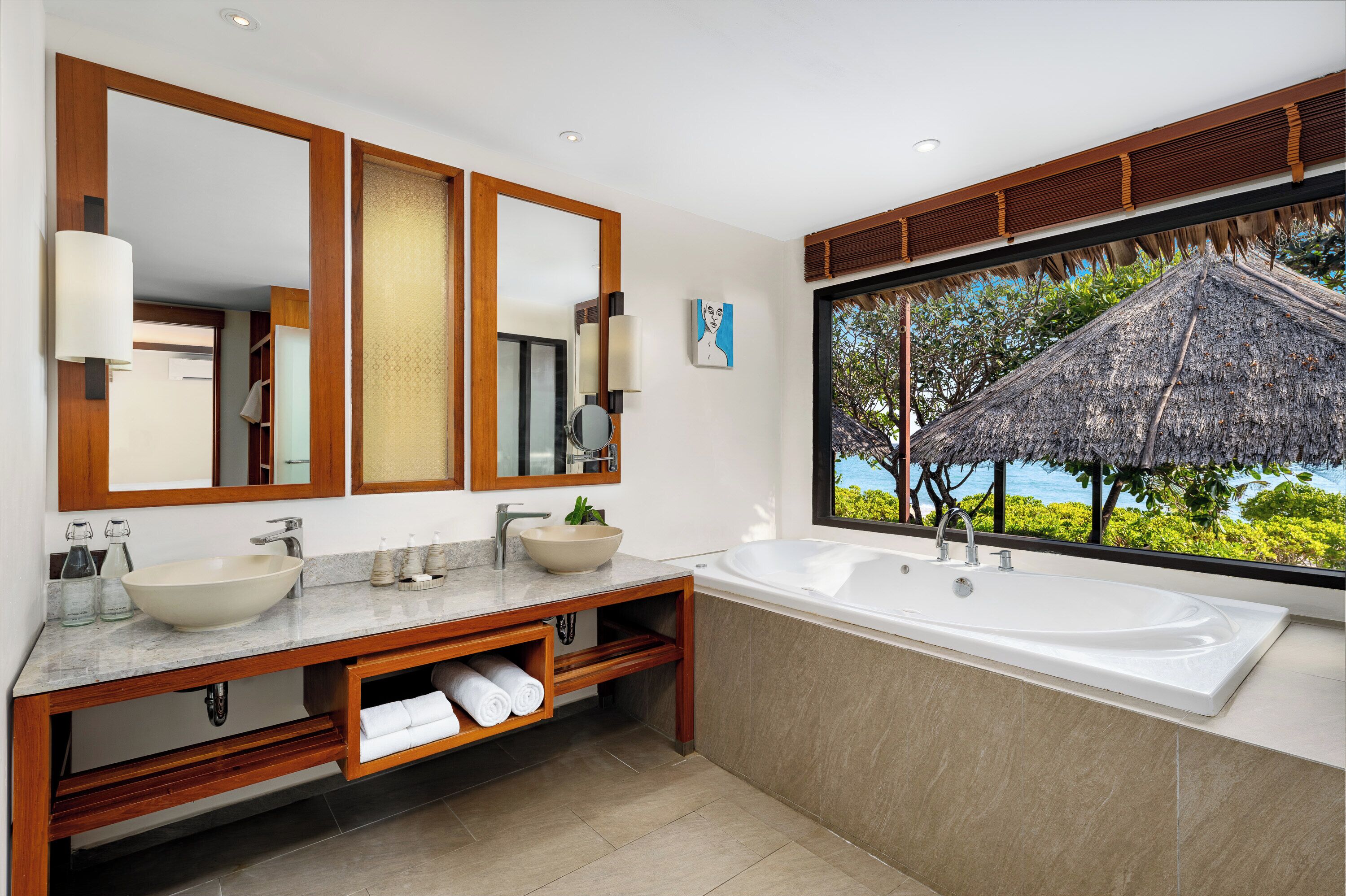 A modern bathroom with a large mirror, two sinks, a bathtub, and a view of a thatched-roof structure.