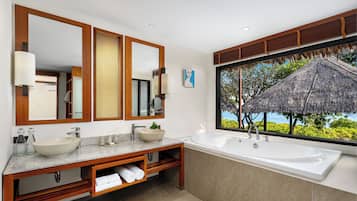 A modern bathroom with a large mirror, two sinks, a bathtub, and a view of a thatched-roof structure.