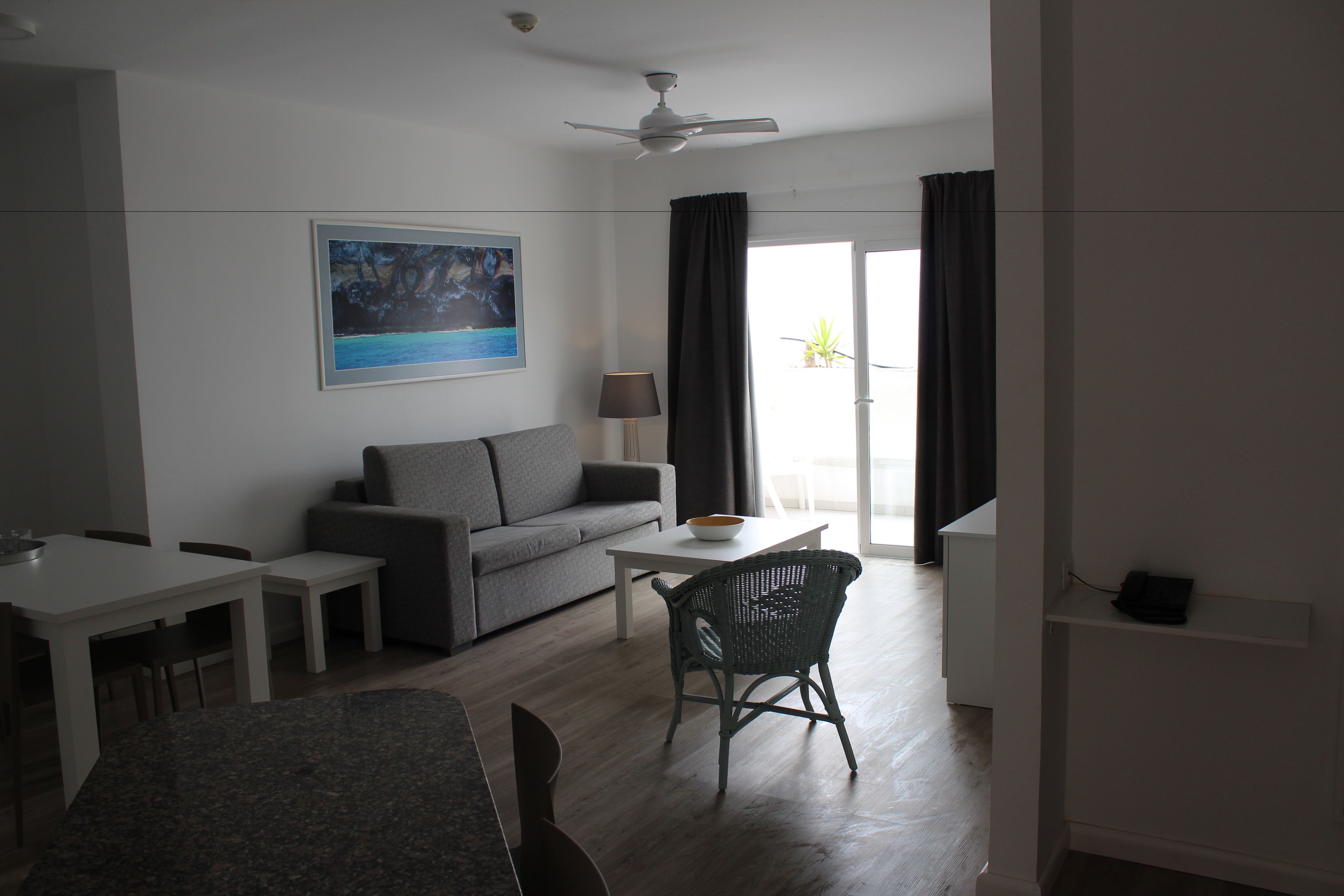 Ein modernes Wohnzimmer mit einer Couch, einem Esstisch und Stühlen. Es gibt einen Deckenventilator, ein Bild an der Wand und einen Blick ins Freie durch ein Fenster.
