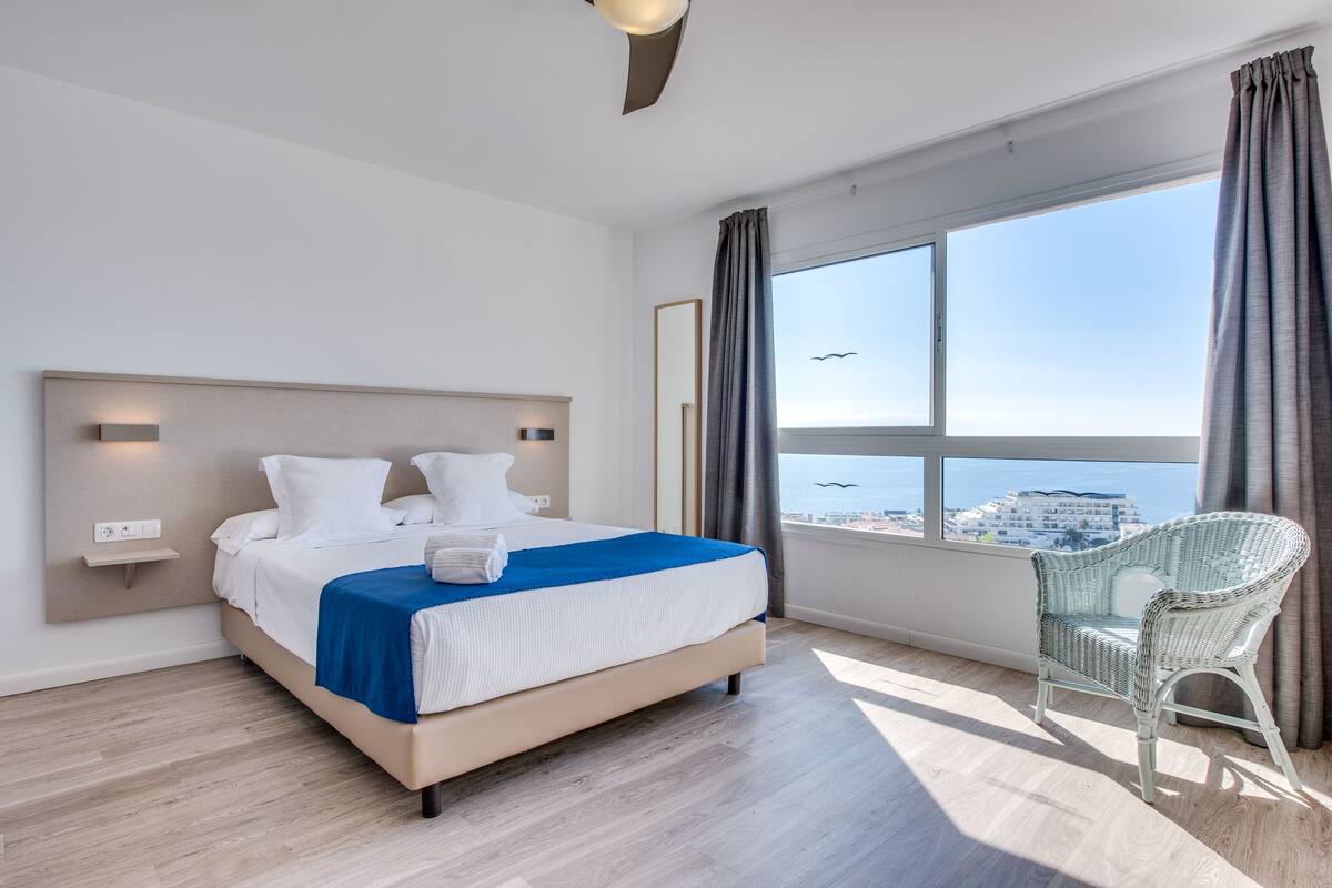 Ett modernt hotellrum med en stor säng, utsikt över havet och en fåtölj i rotting.