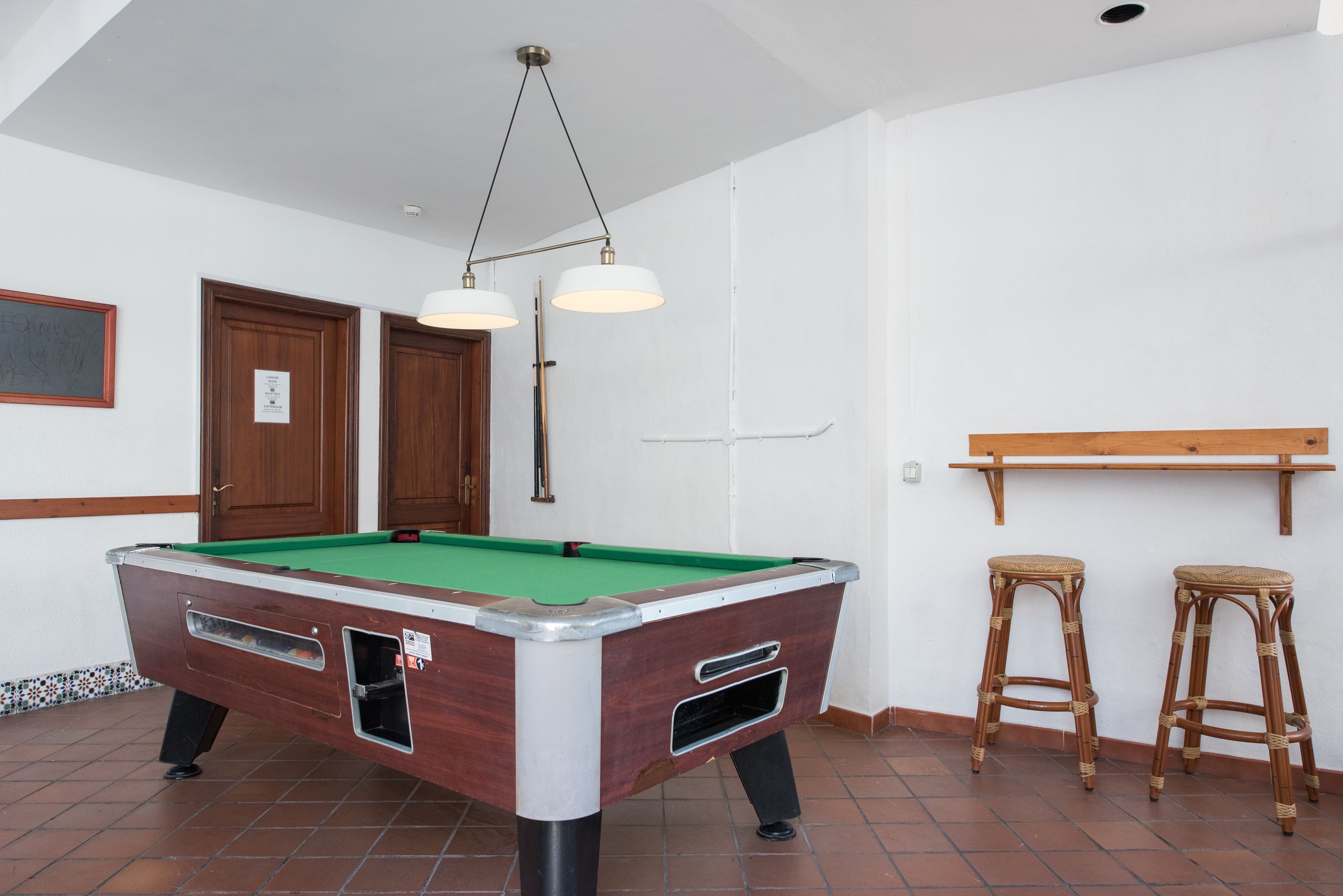 Una mesa de billar con superficie de fieltro verde, dos taburetes de madera y una lámpara colgante.