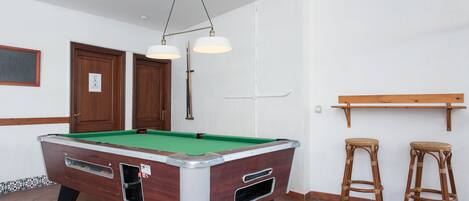 Una mesa de billar con superficie de fieltro verde, dos taburetes de madera y una lámpara colgante.