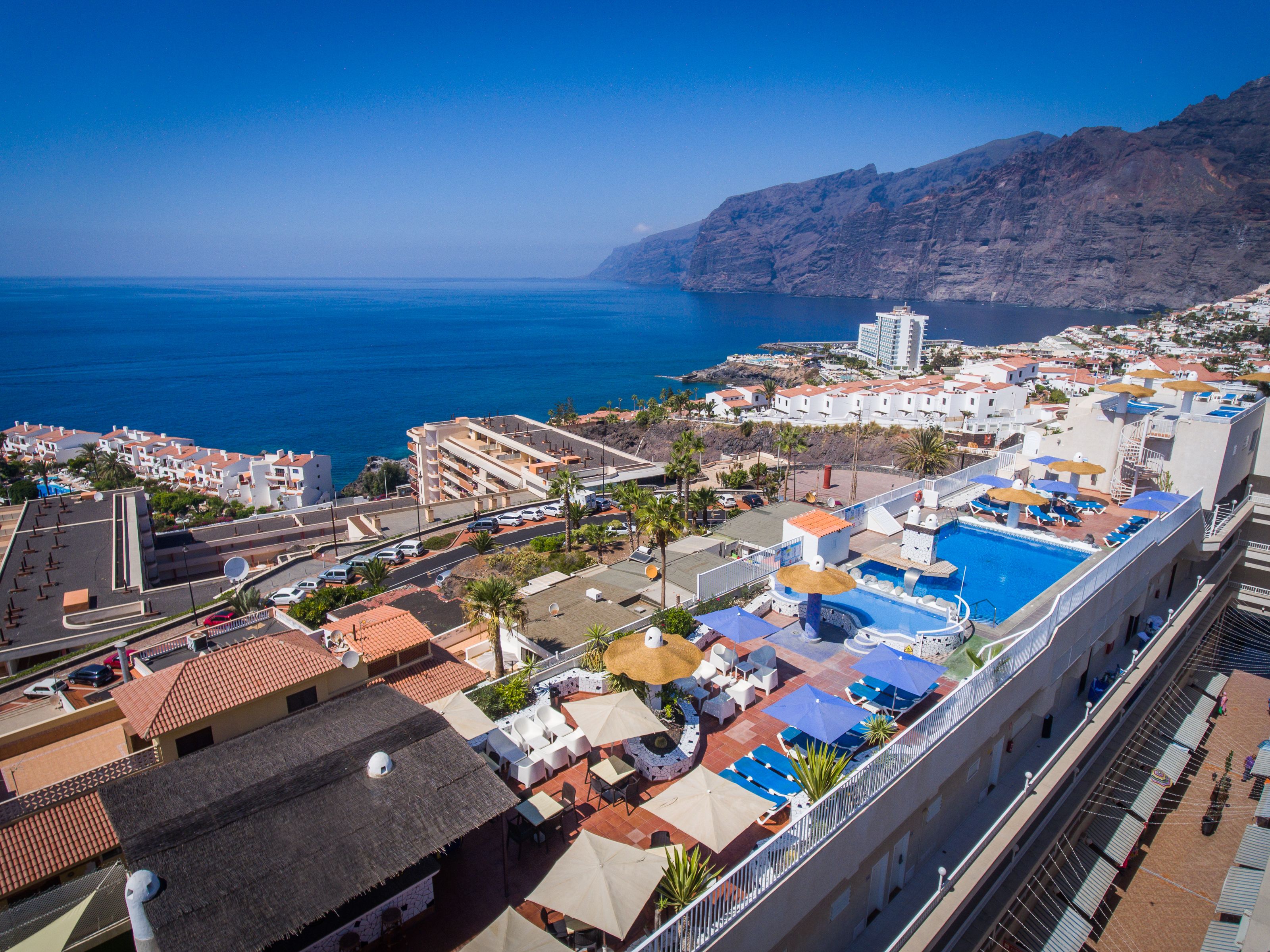 Un hotel frente al mar con piscina en la azotea, varios balcones y vistas al mar y a las montañas.