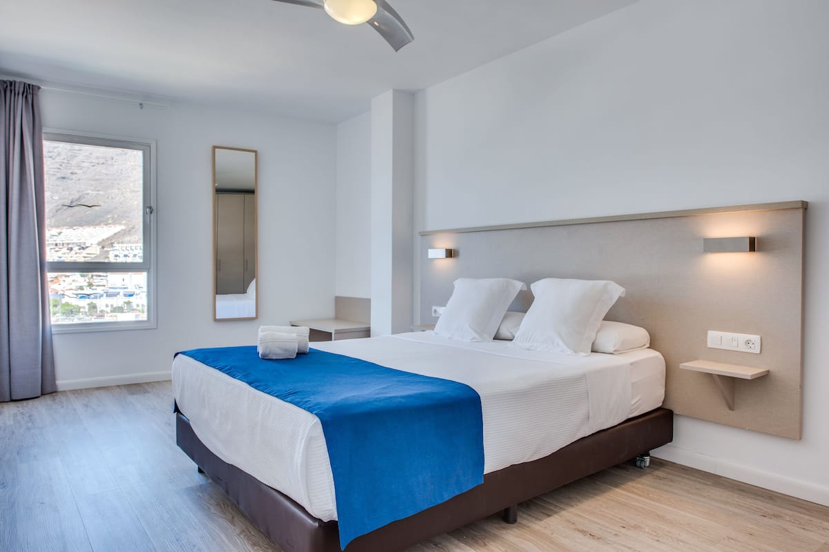 Ett modernt hotellrum med en stor säng, ett sängbord, en liten avlastningsyta, en spegel och utsikt över staden genom ett fönster.