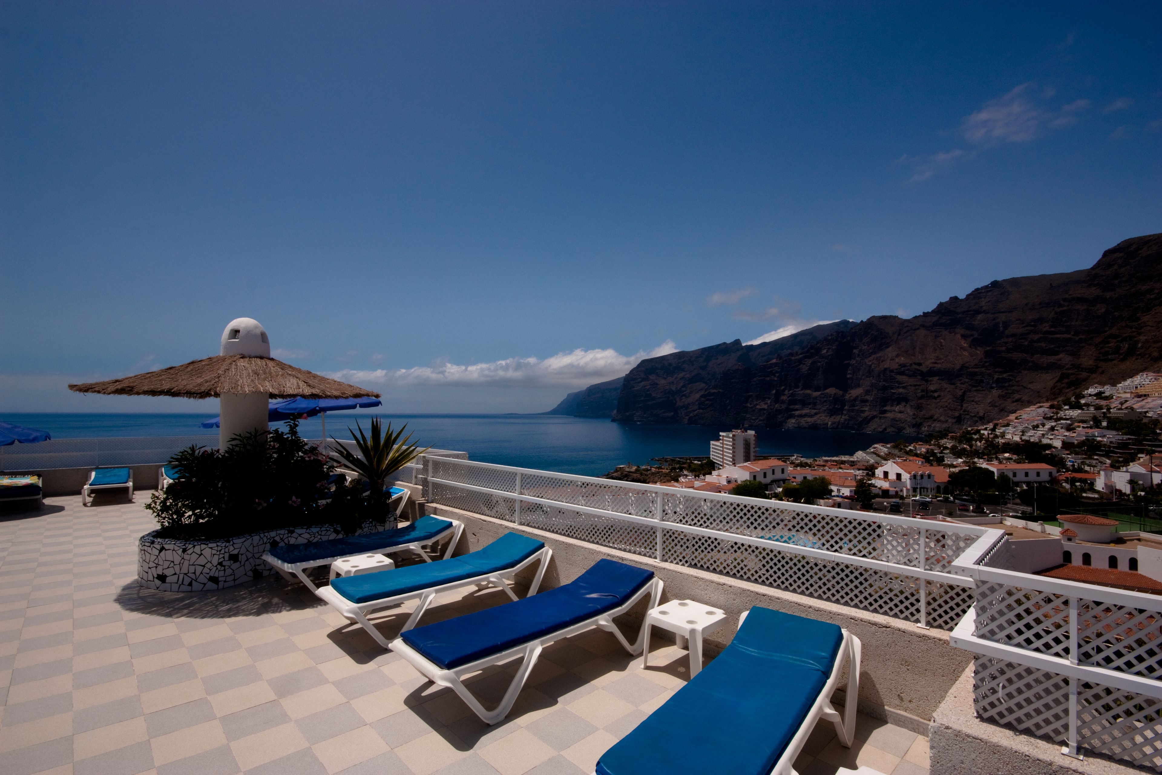A terrace with lounge chairs and a thatched umbrella, overlooking a coastal town and the sea.