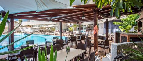 Ein Außenpoolbereich mit Liegestühlen und Tischen, umgeben von viel Grün und beschattet durch eine Pergola.