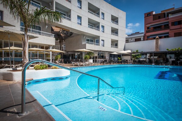 Une piscine à l’eau d’un bleu limpide, entourée de chaises longues et de parasols, avec un immeuble à plusieurs étages en arrière-plan.