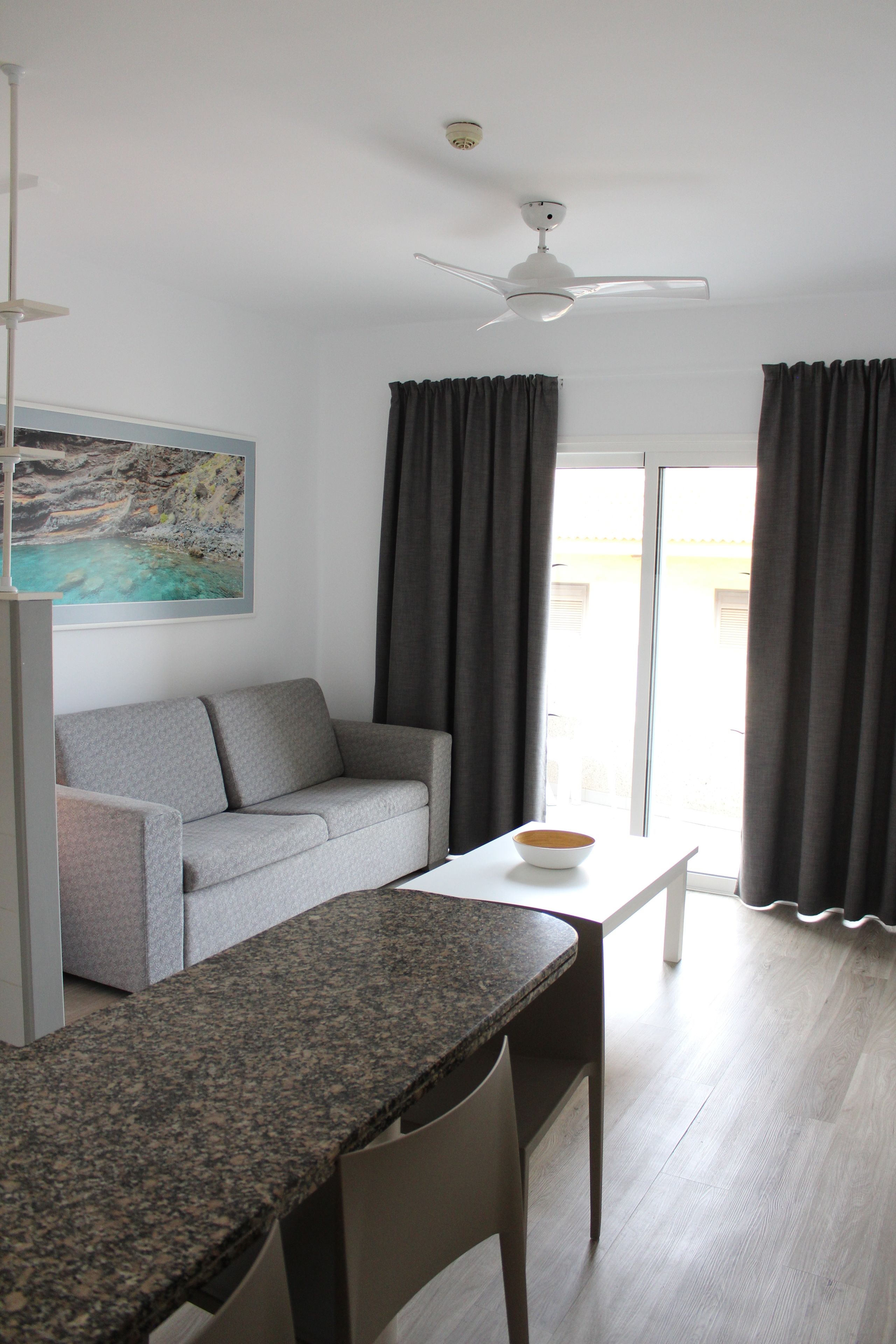 Ein modernes Wohnzimmer mit grauem Sofa, einem kleinen Tisch mit Schale, Deckenventilator und einem großen Fenster mit Vorhängen.