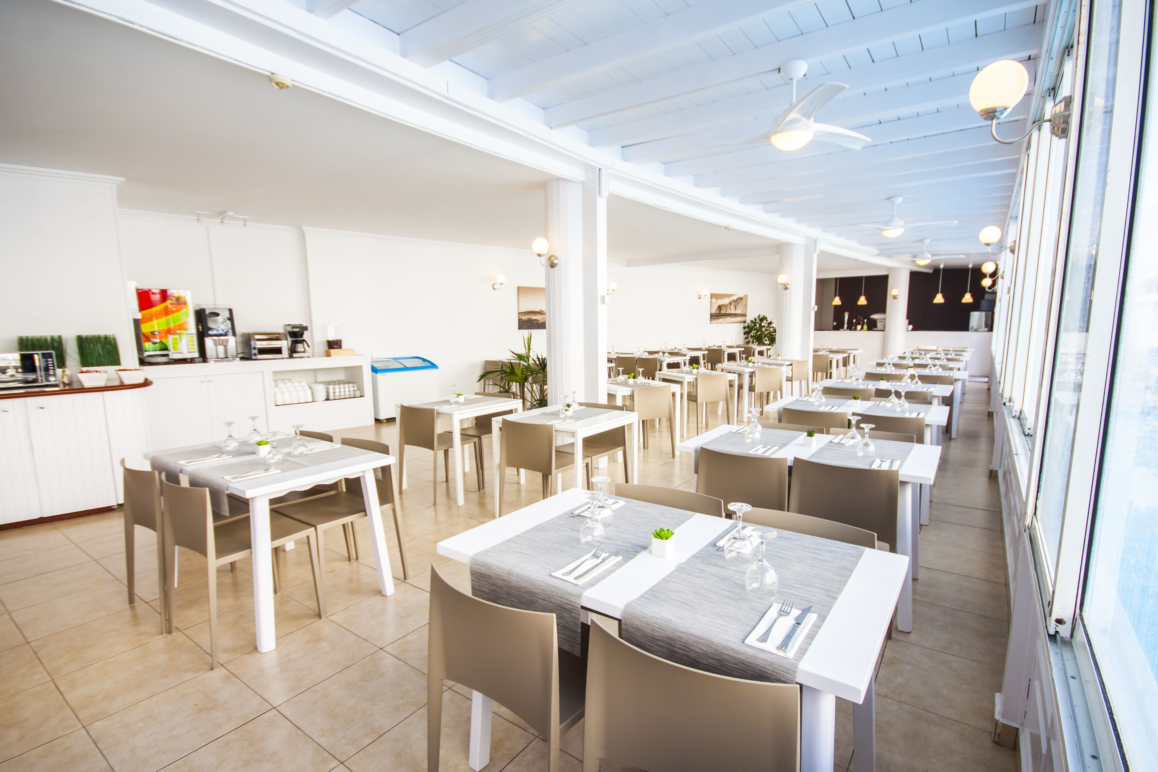 A spacious dining area with multiple tables and chairs, a kitchen area with appliances, and a large window providing natural light.