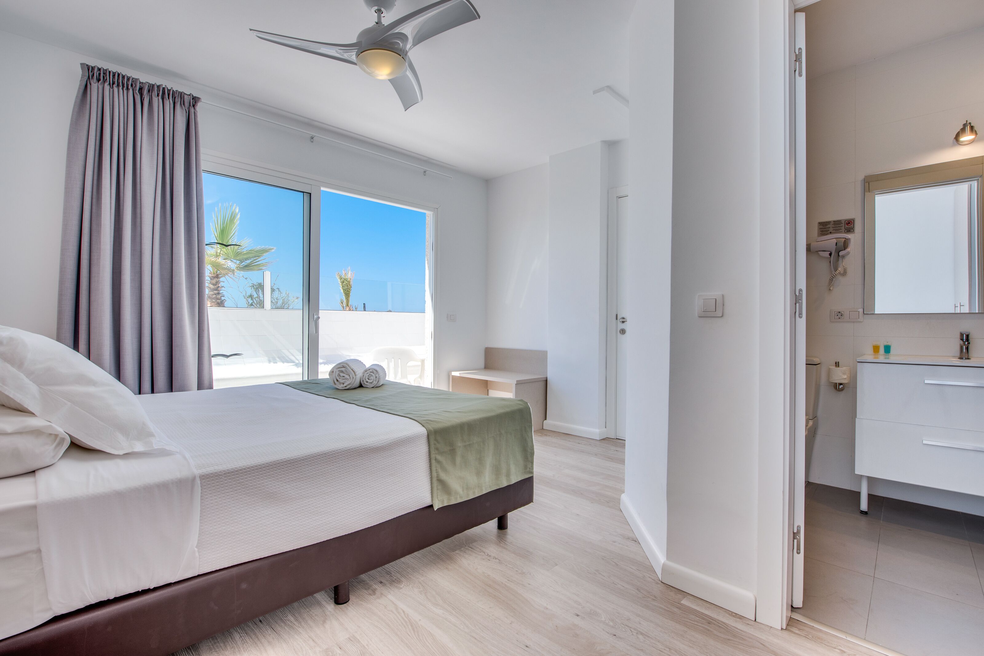 A bedroom with a bed, a ceiling fan, a view of the outdoors, a bathroom with a mirror and sink, and a door leading to another room.