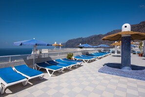Un espace extérieur ensoleillé, aménagé avec des fauteuils bleus et des parasols, offrant une vue sur un paysage côtier.