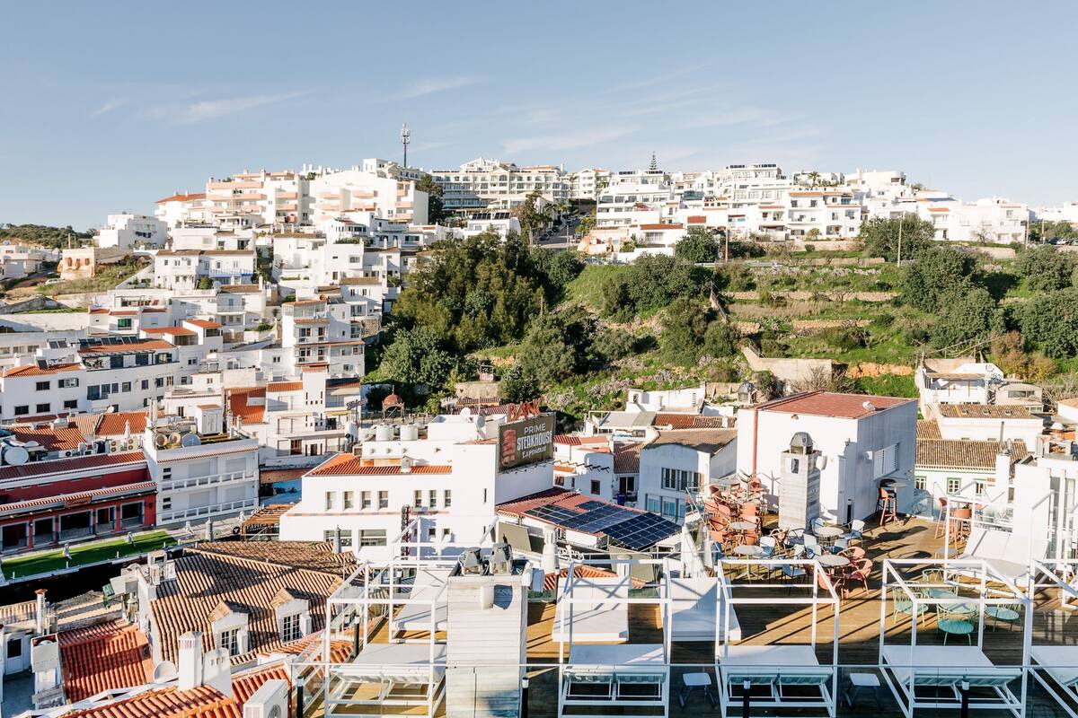A coastal town with white buildings, a hill with greenery, and a rooftop bar with outdoor seating.