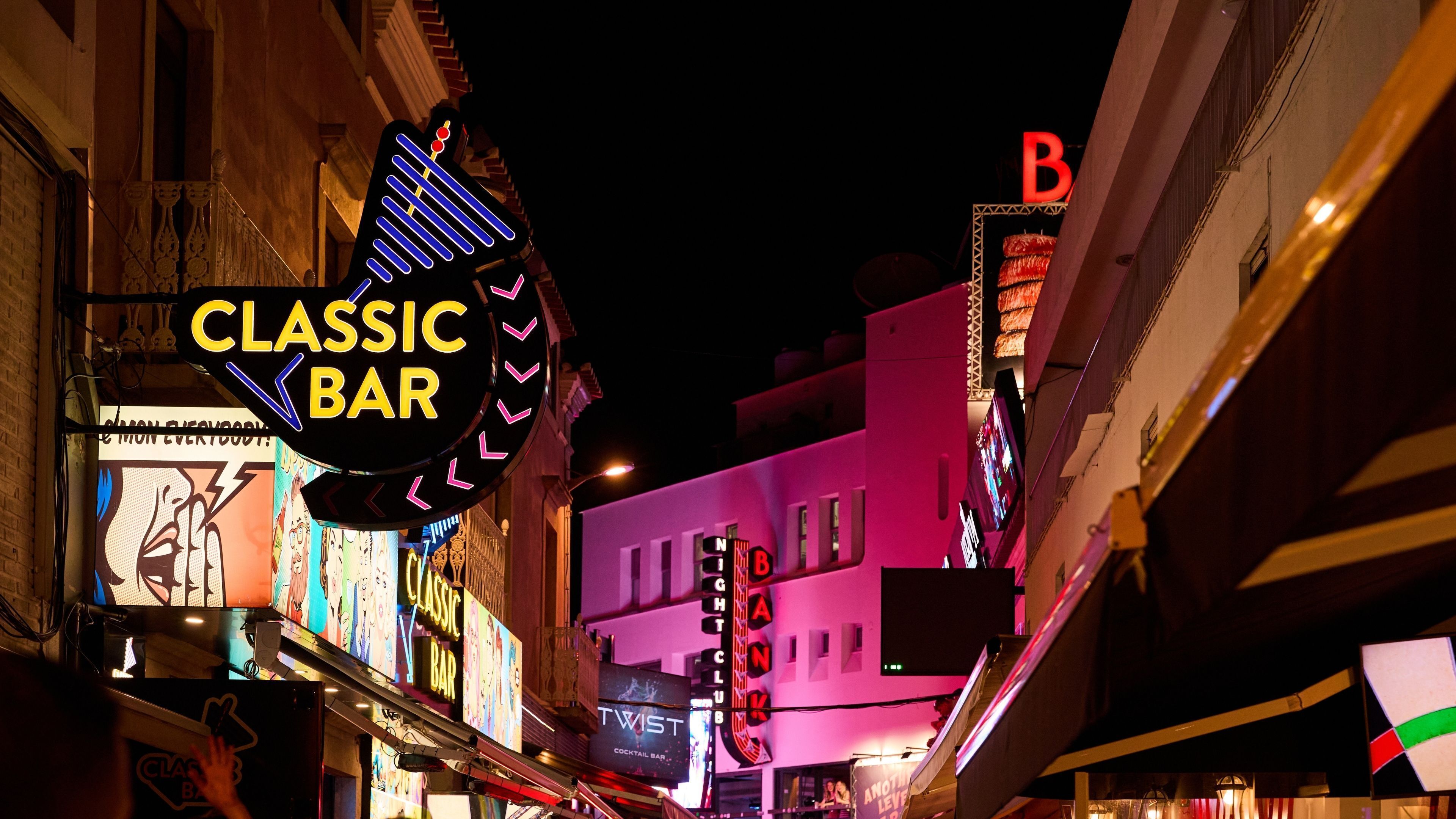 Eine klassische Bar mit Neonreklamen und einer Neon-Gitarre, gelegen in einer schmalen Straße, gesäumt von Gebäuden.