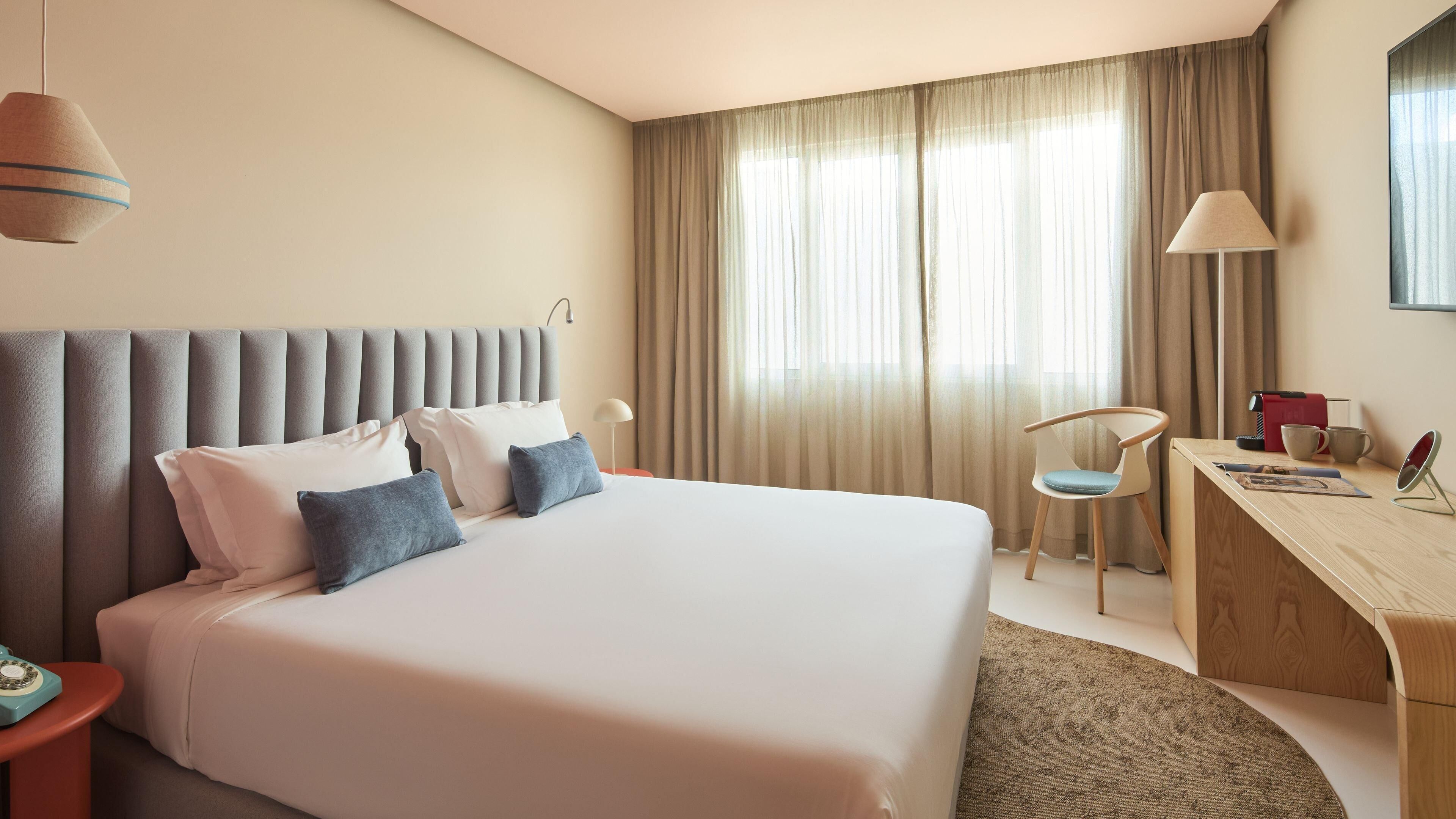 A hotel room with a large bed, a desk, a chair, and a window with curtains.