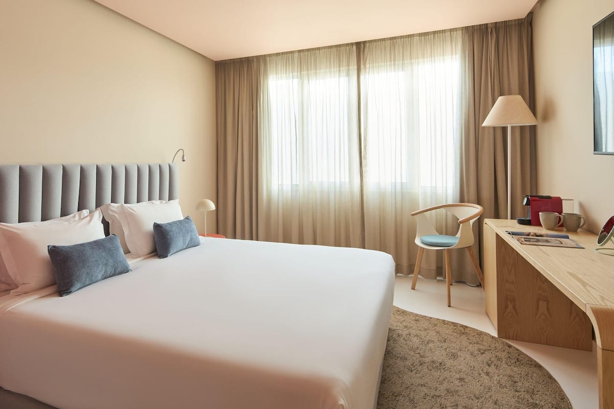 A hotel room with a large bed, a desk, a chair, and a window with curtains.