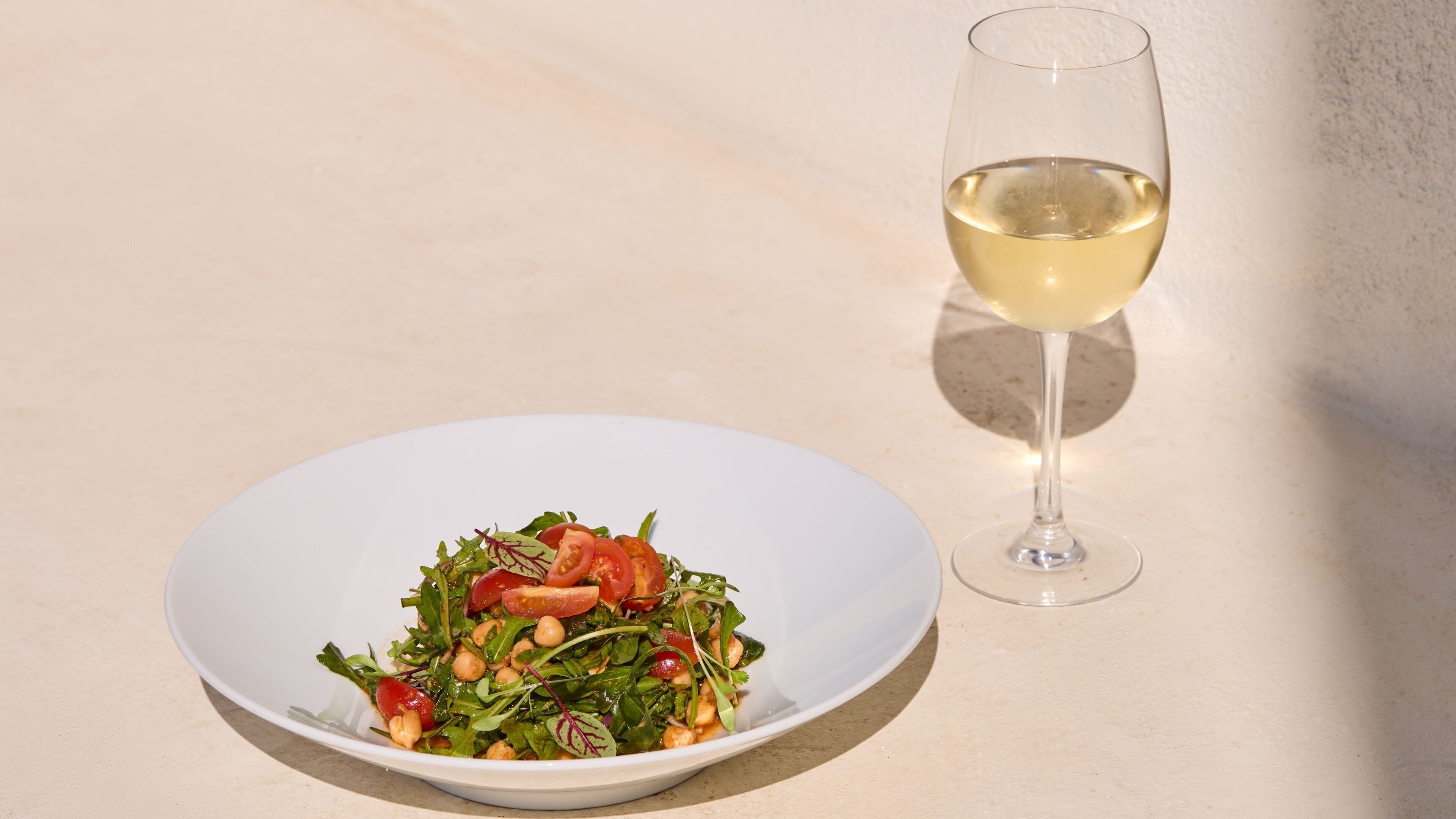 A white plate with a salad and a glass of white wine on a light-colored surface.