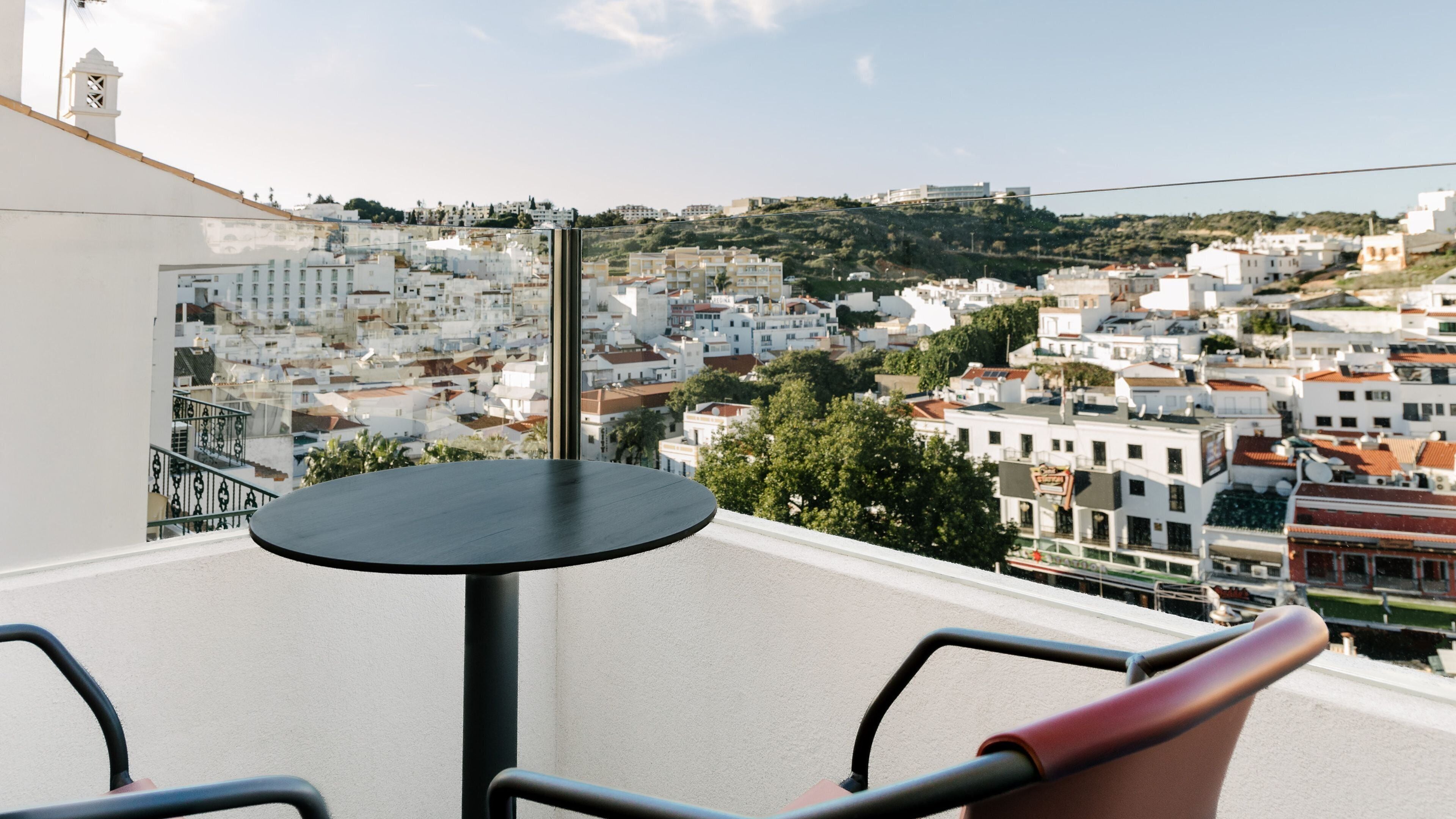 A balcony with a round table and chairs, overlooking a densely packed town with white buildings and a hill in the distance.