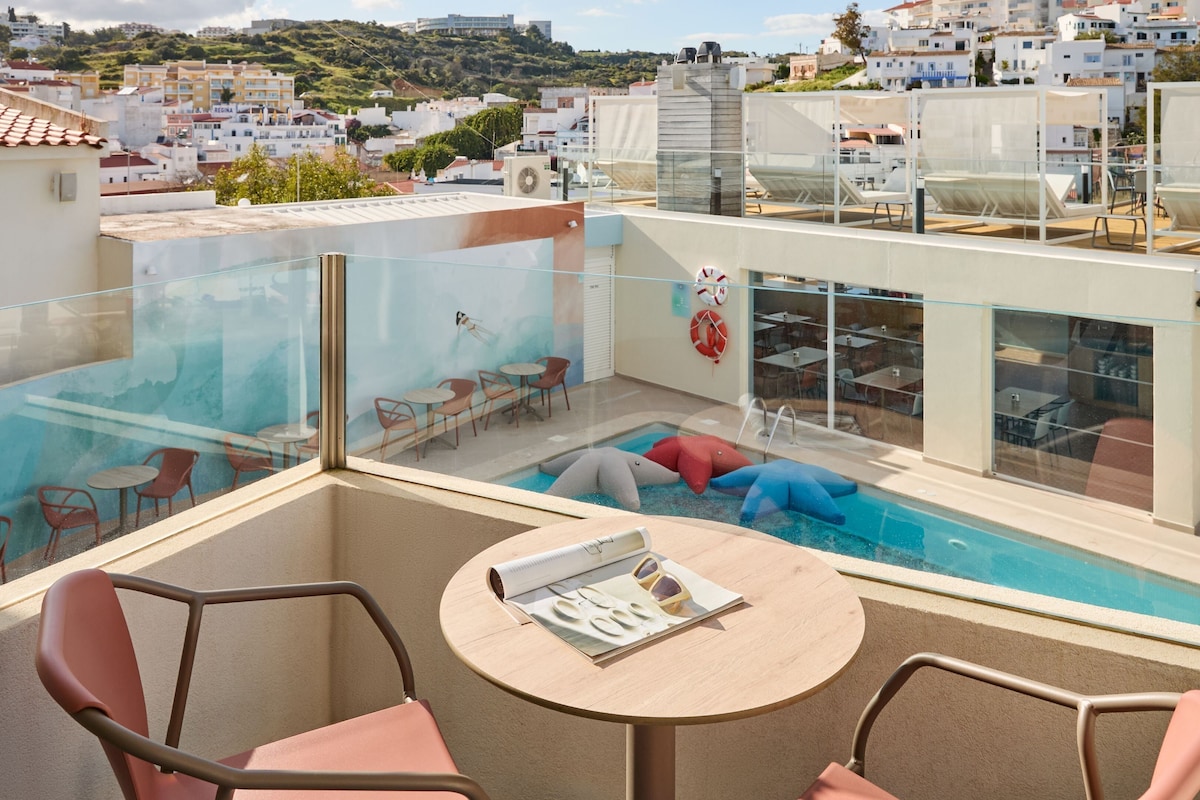 Un área de piscina en la azotea con barandilla de cristal, mobiliario de piscina y vistas de la ciudad.