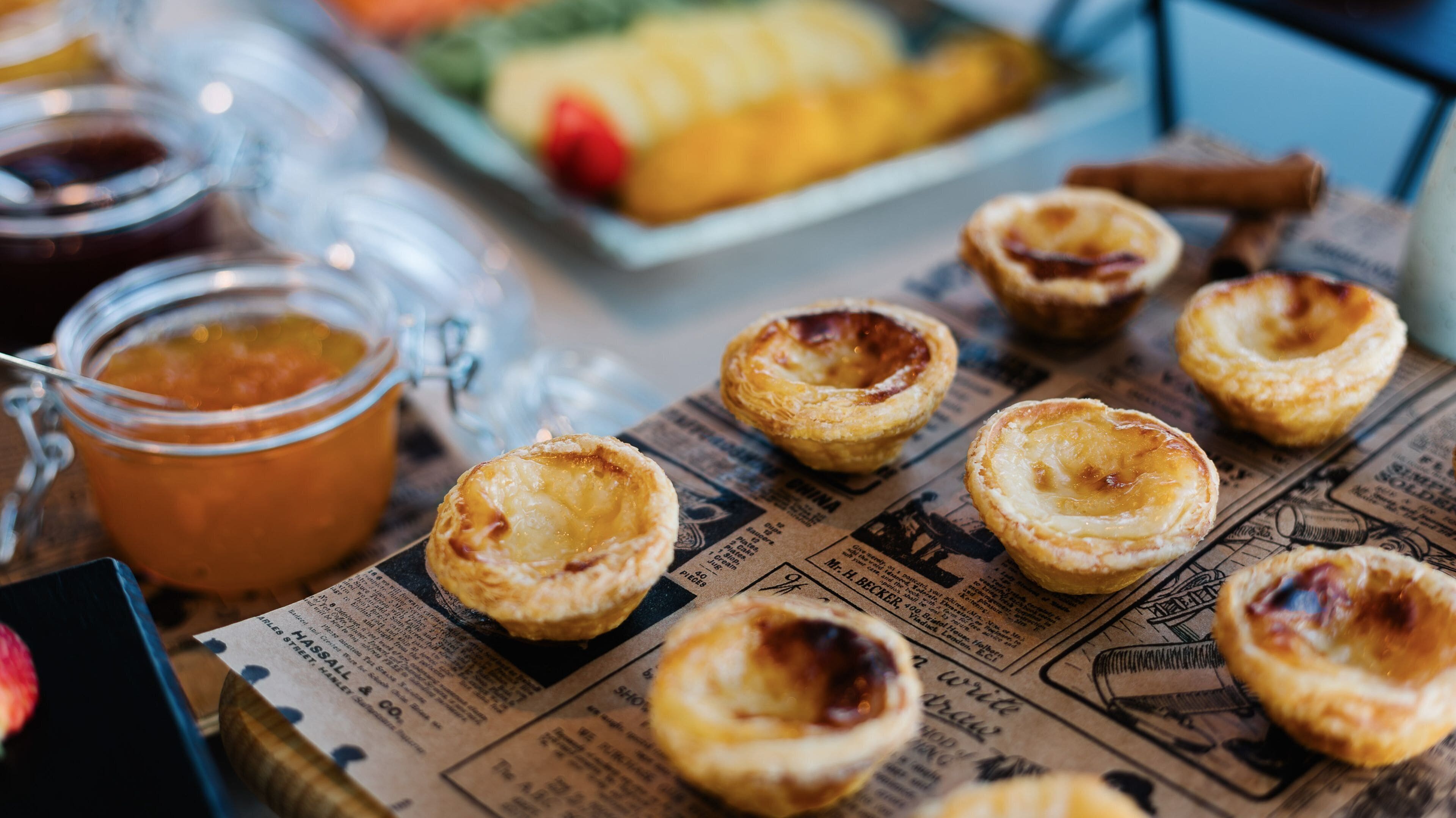 A table with several small pastries, a glass jar of jam, and a newspaper.