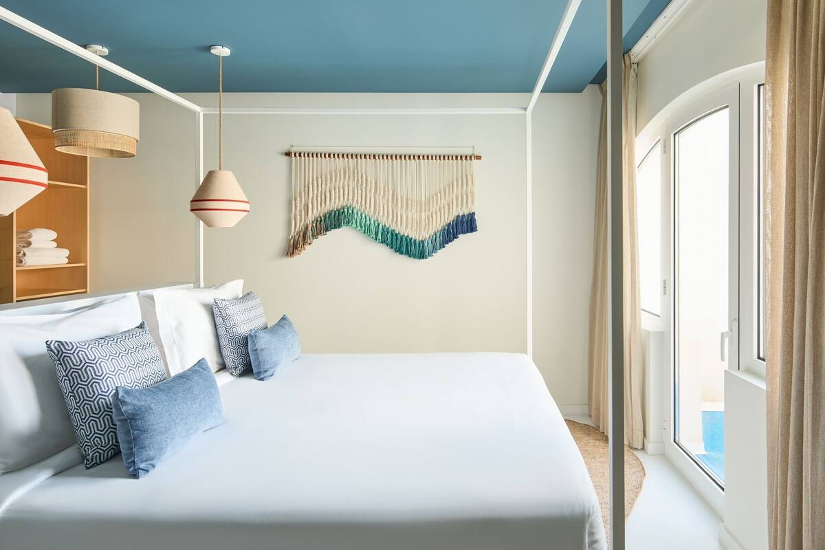 A modern hotel room with a large bed, hanging pendant lights, a woven wall hanging, and a view of the sea through a large window.