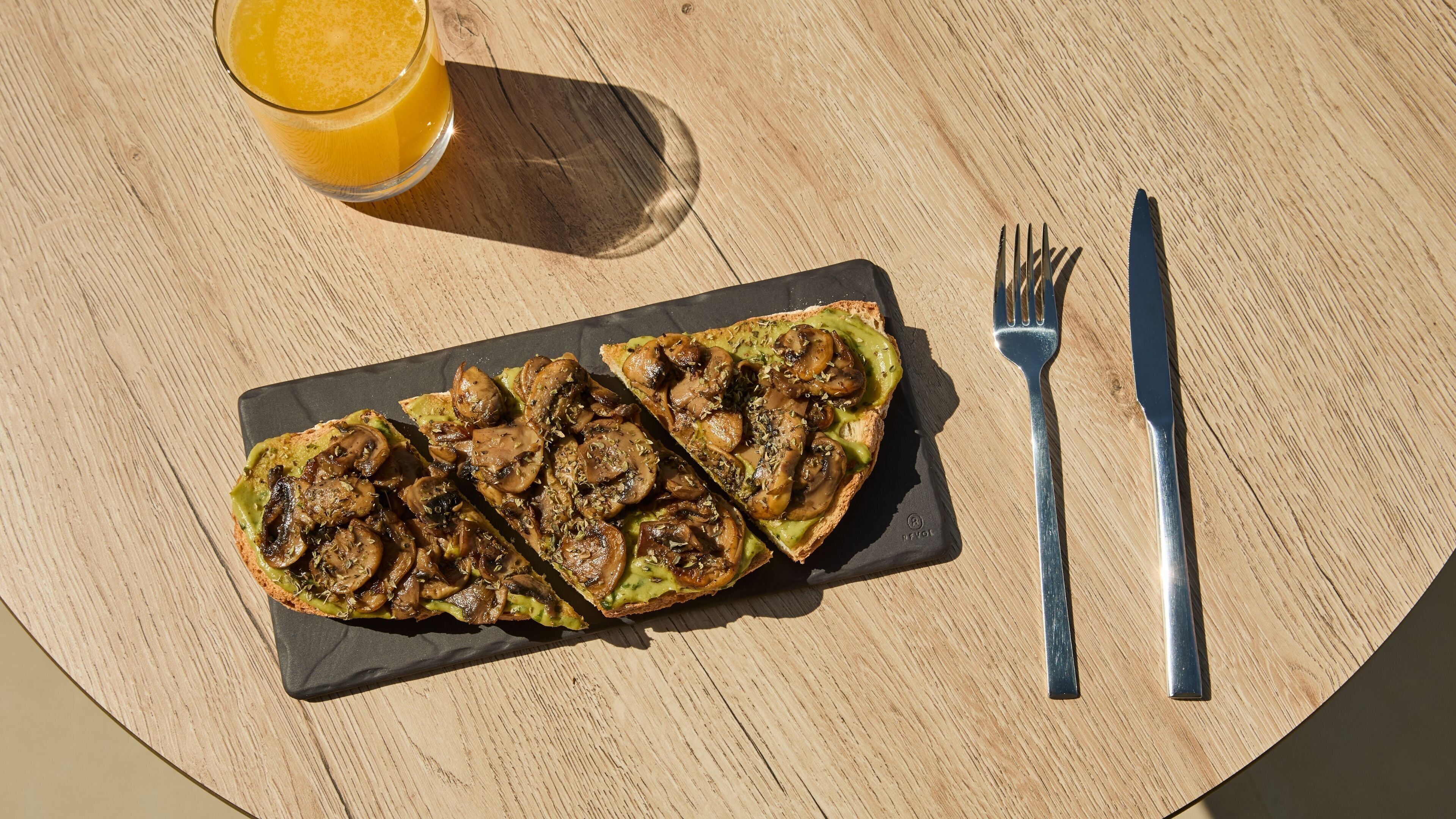 Een maaltijd bestaande uit een gegrilde schotel met paddenstoelen, geserveerd met een glas sinaasappelsap, op een ronde houten tafel.