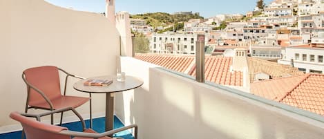 Un balcon avec deux chaises et une petite table, d’où l’on peut admirer une colline parsemée de bâtiments blancs aux toits de tuiles rouges.