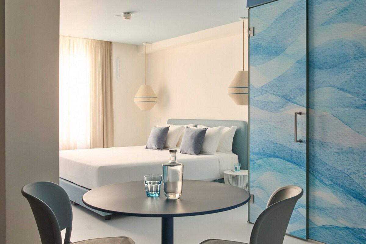 A modern hotel room with a bed, a round table with chairs, a bottle of water, and a glass.