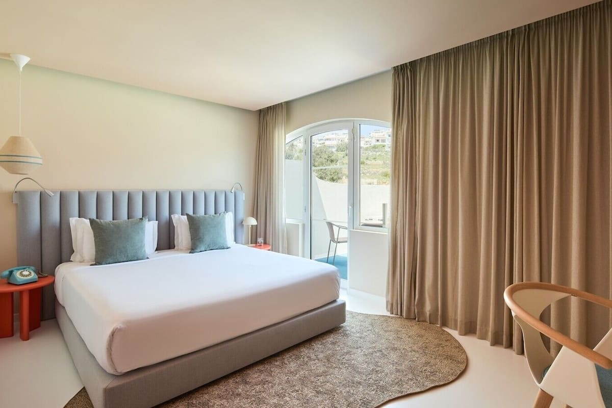 Habitación de hotel con una cama grande, una mesita de noche, una lámpara, una silla y vistas al exterior.