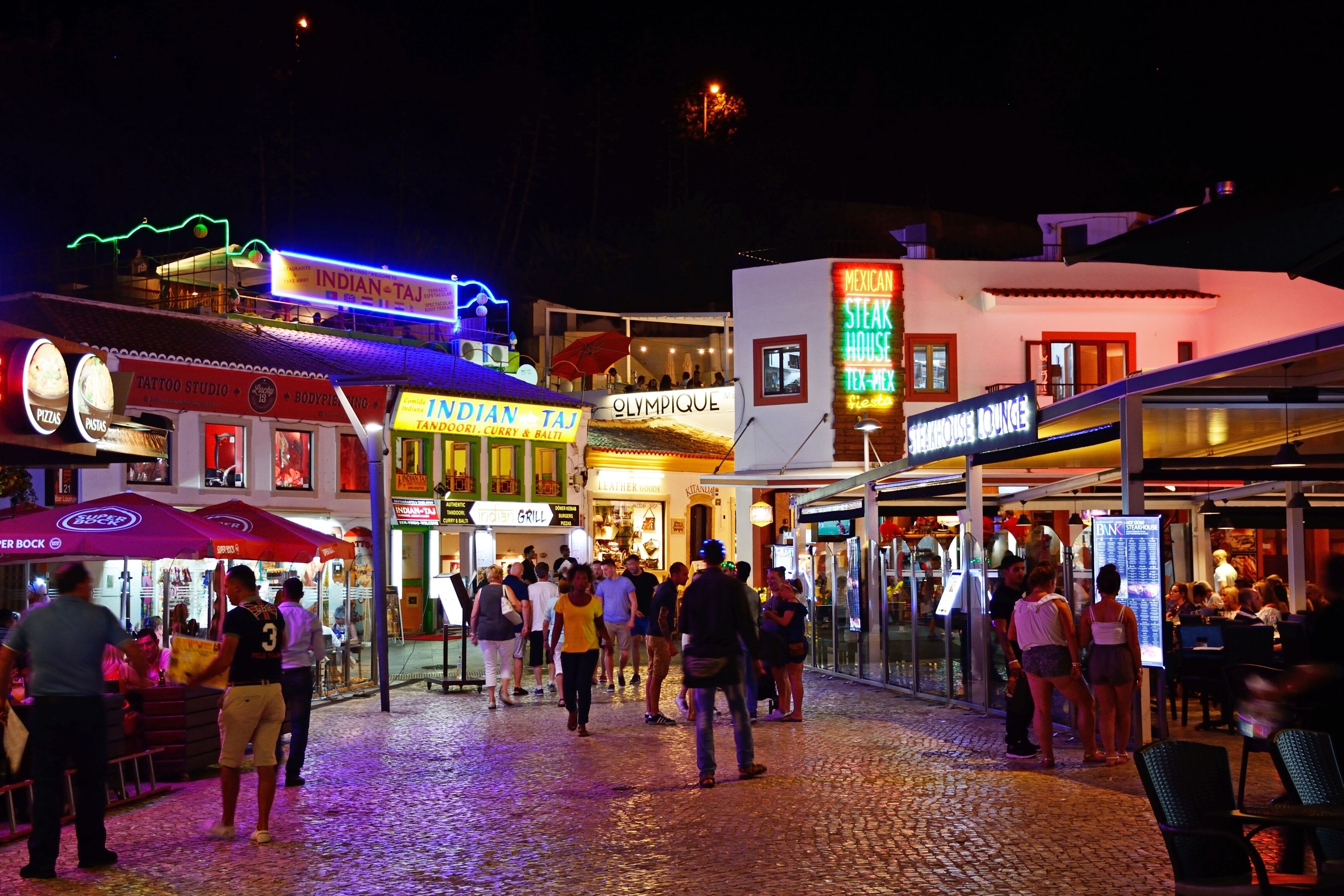 A bustling street scene at night with various shops and restaurants, including an Indian Taj and a steak house.