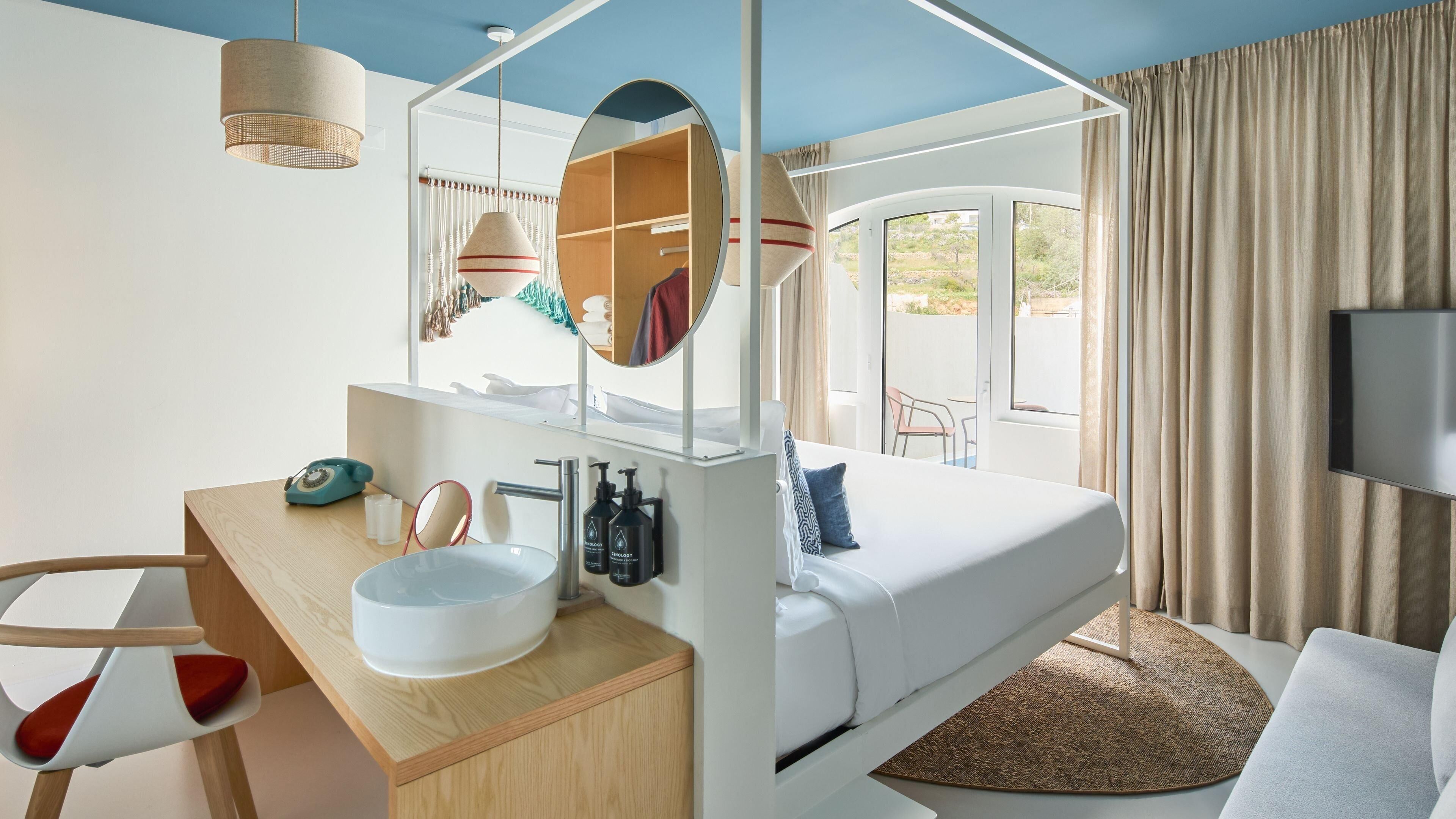 A modern hotel room with a bed, a desk with a sink, a mirror, and a TV.