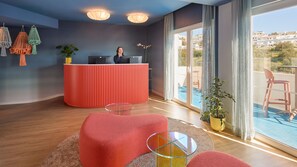 A modern hotel lobby with a red reception desk, a large window with a view of the city, and a seating area with round tables and chairs.