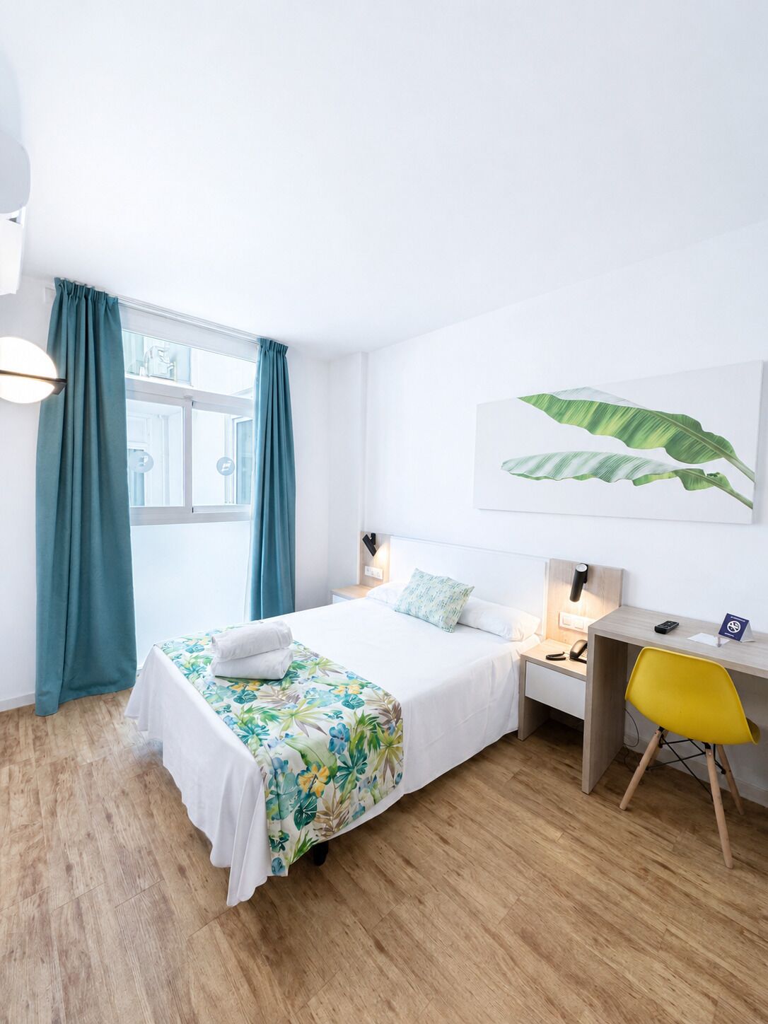 Habitación de hotel con cama, escritorio, silla, ventana con cortinas y una obra de arte en la pared.