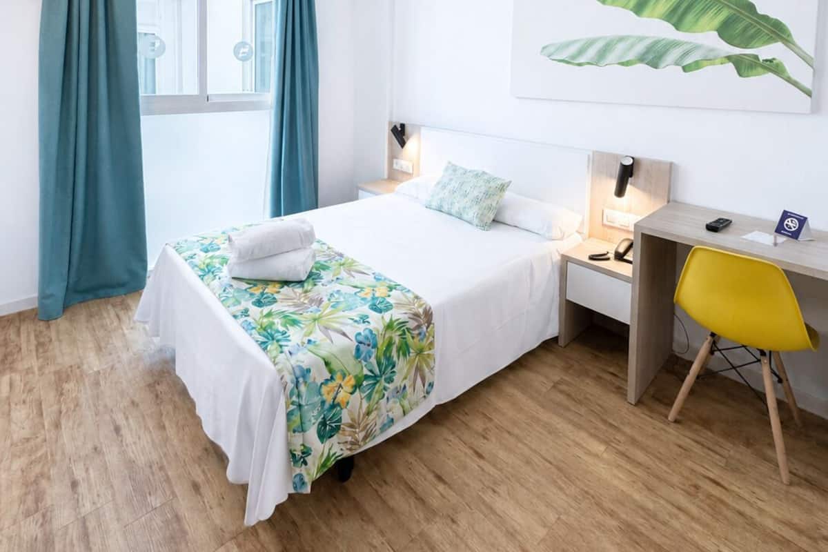 Habitación de hotel con cama, escritorio, silla, ventana con cortinas y una obra de arte en la pared.