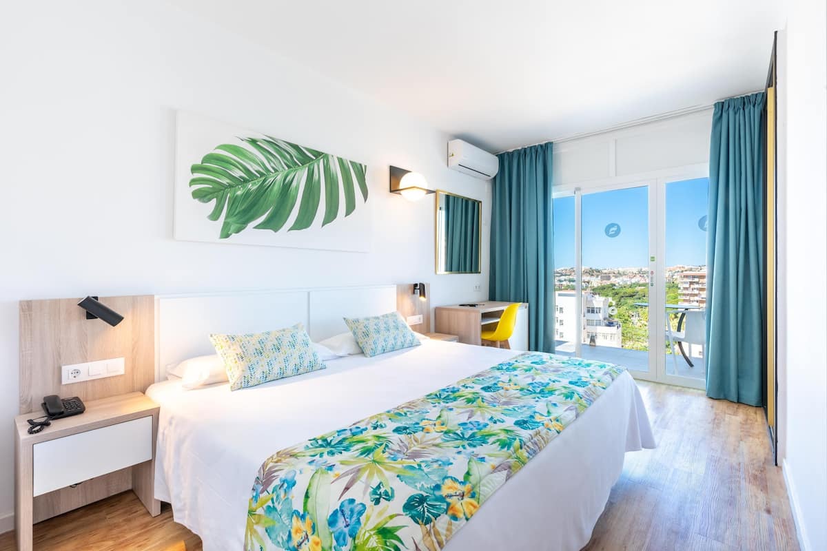 A hotel room with a bed, bedside table, lamp, and a view of the city through large windows.