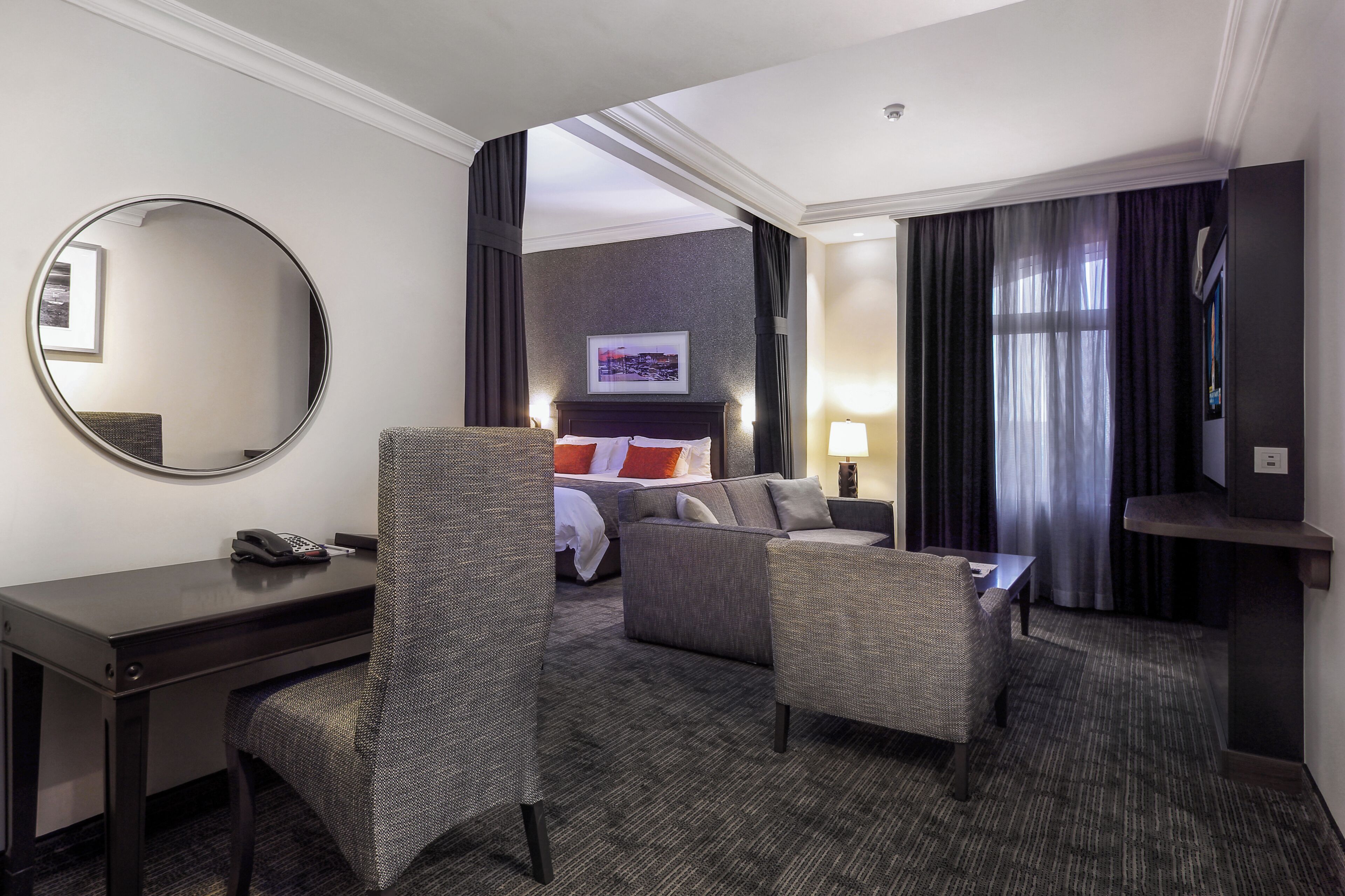 A hotel room with a bed, two armchairs, a desk, a mirror, a TV, and a window with curtains.