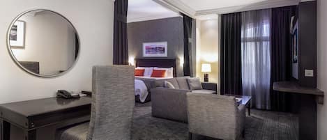 A hotel room with a bed, two armchairs, a desk, a mirror, a TV, and a window with curtains.
