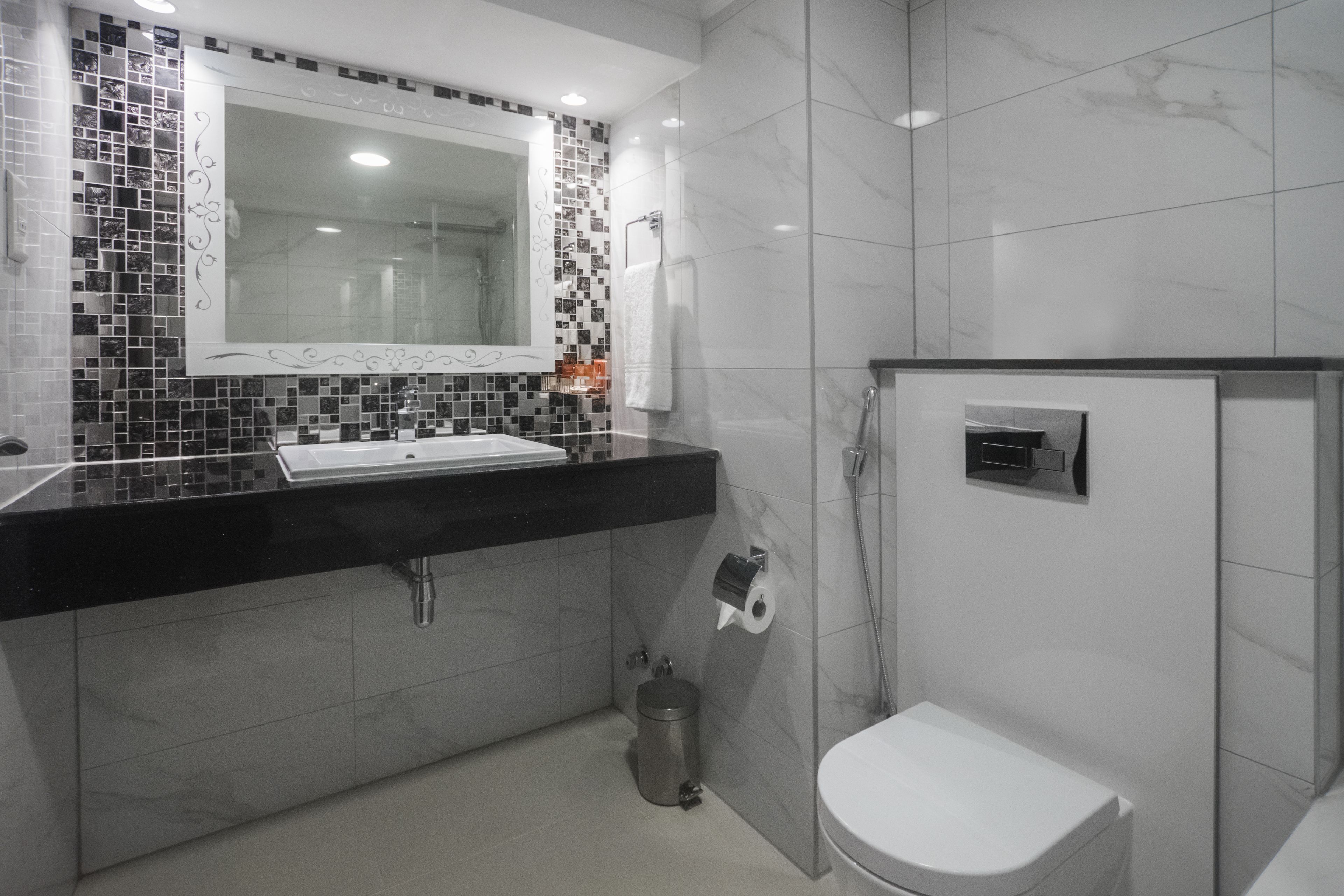 A modern bathroom with a black countertop, a white sink, a mirror with a mosaic backsplash, a toilet, and a shower area.
