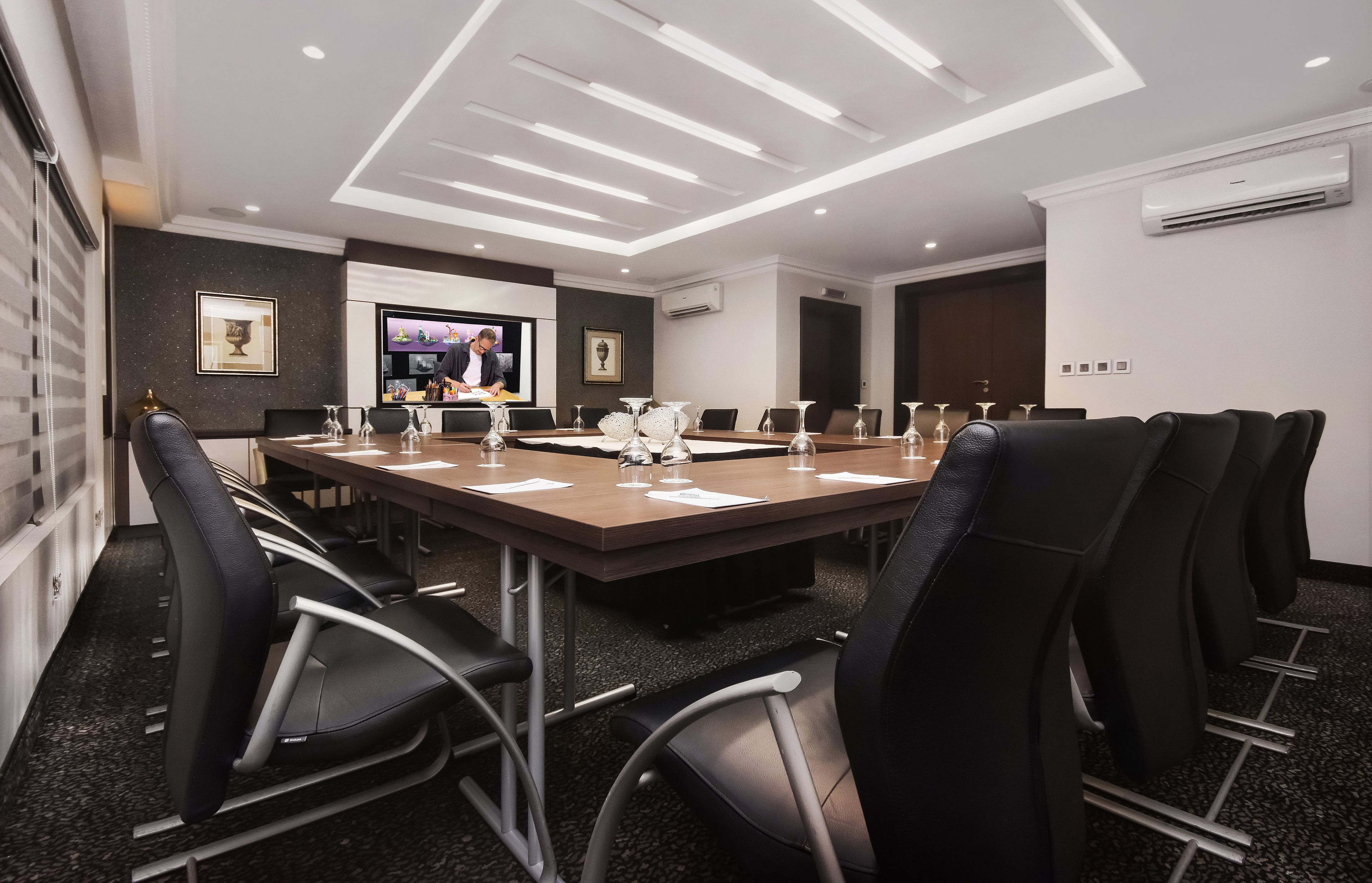 Une salle de conférence avec une longue table, des chaises noires et un écran de télévision.