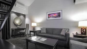 Una habitación de hotel moderna con un sofá gris, un espejo redondo, una mesa de comedor y una fotografía de un paisaje urbano en la pared.