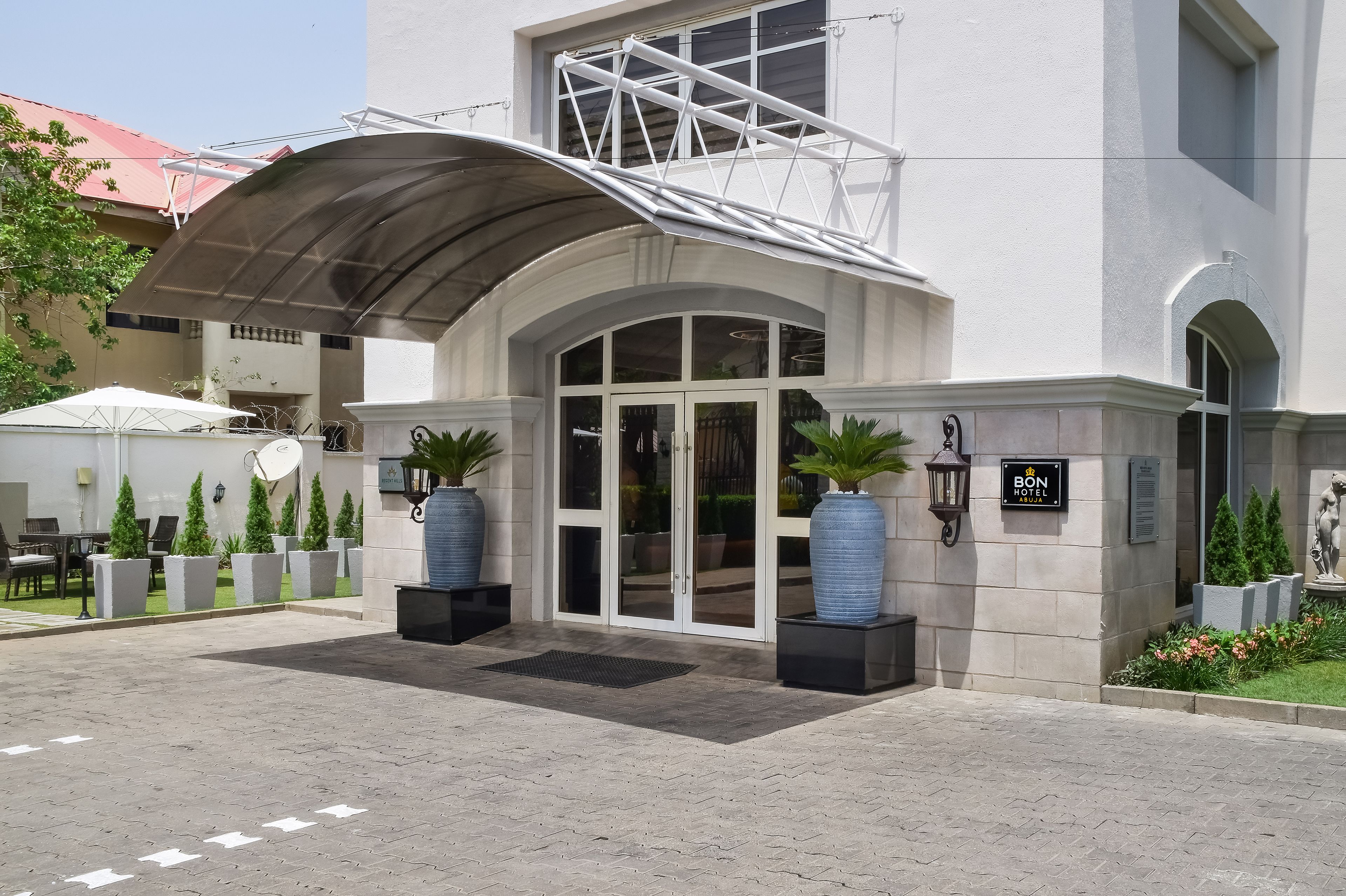 L’entrée d’un hôtel, avec un passage couvert, de grands pots de plantes et une enseigne indiquant le nom de l’hôtel.