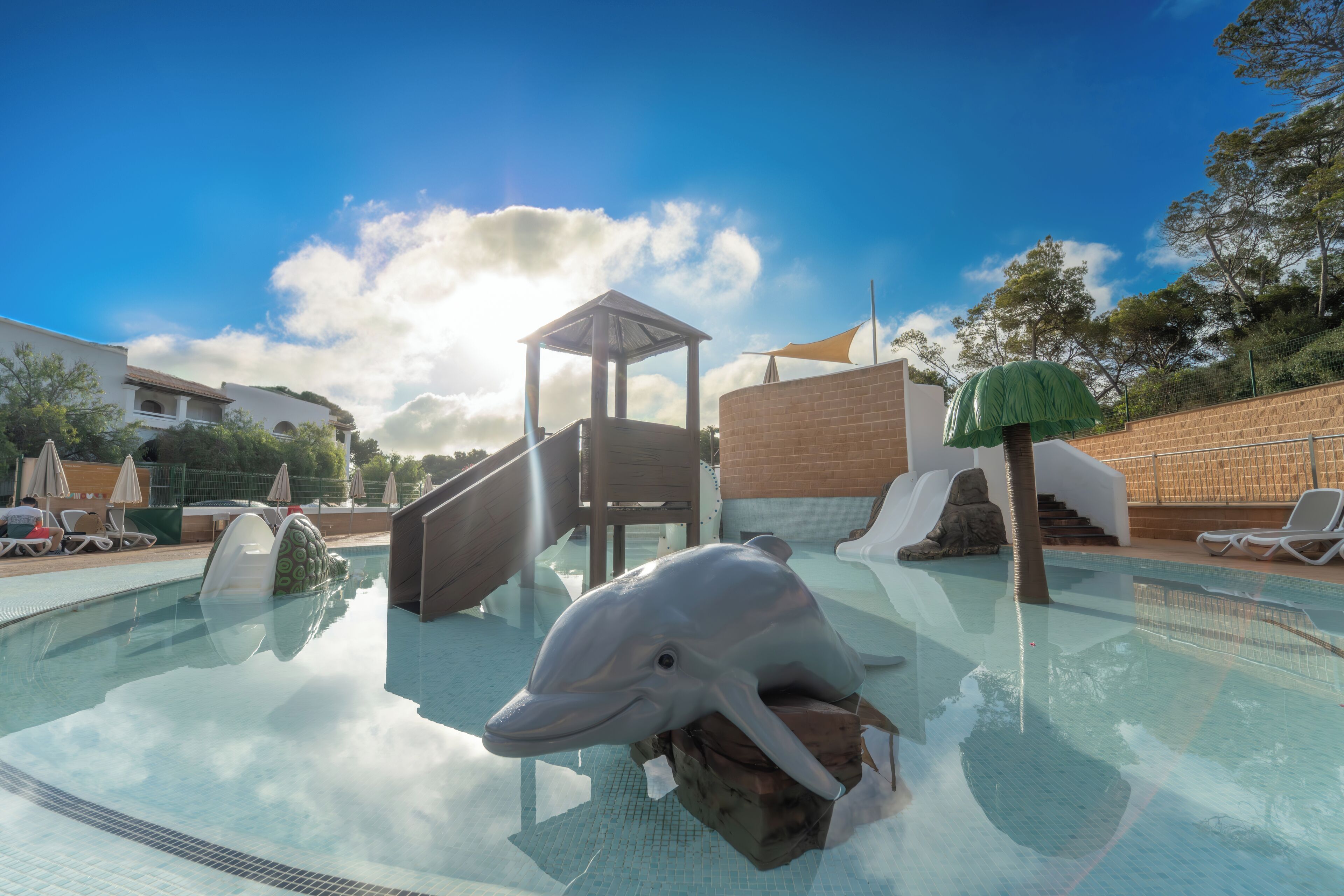 En pool med en delfinskulptur, solstolar och en lekplats.