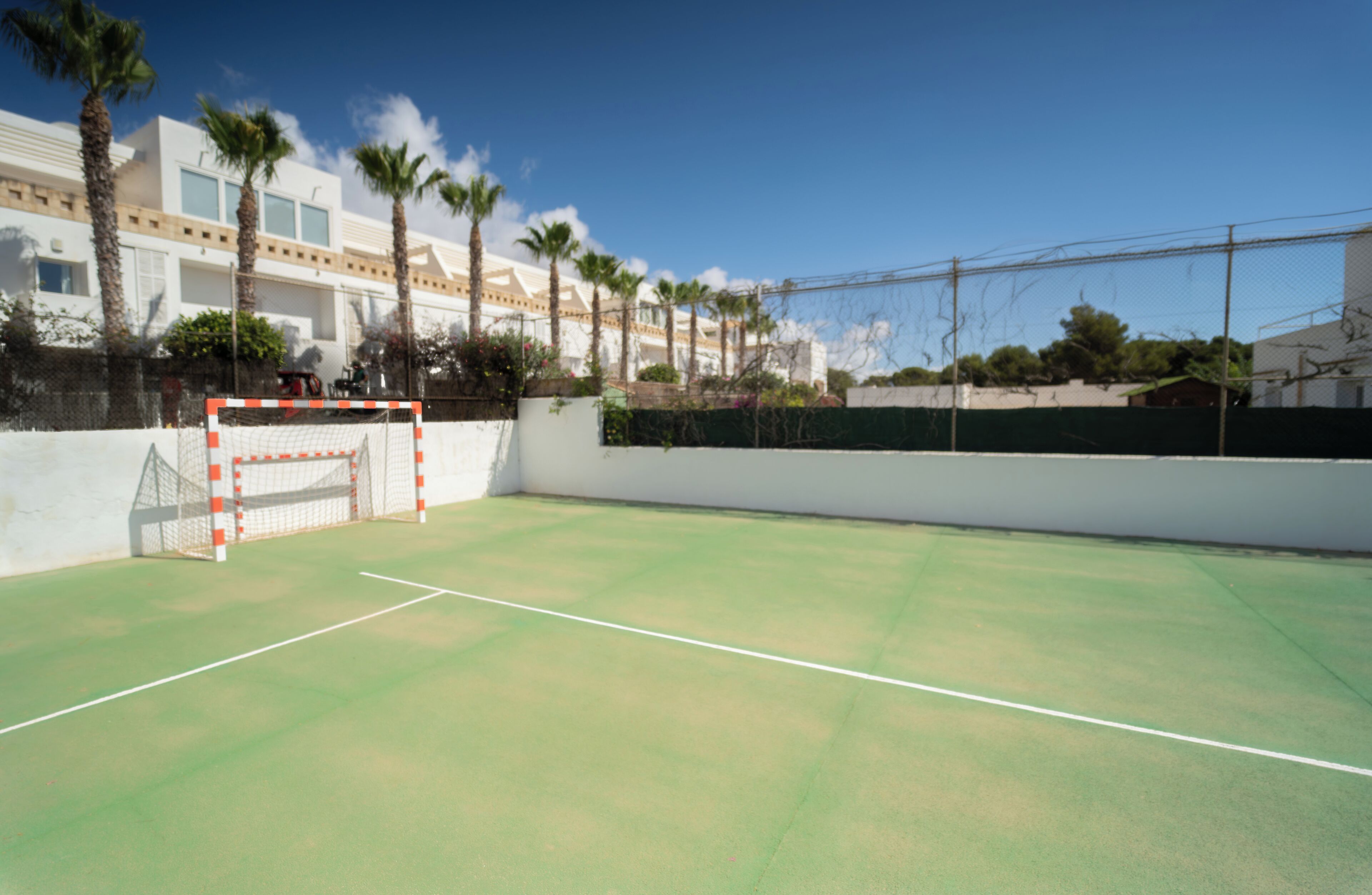 A tennis court with a goal post, palm trees, and a building in the background.