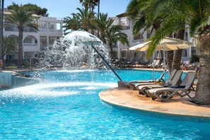 Une piscine agrémentée d’une fontaine, des chaises longues, des palmiers et, au fond, un bâtiment doté de balcons.