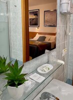 Un bagno con box doccia in vetro, un lavandino bianco con ripiano in vetro, una pianta in vaso e uno specchio che riflette un letto con testiera e due cuscini.