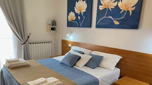 A neatly made bed with a blue and white bedspread, two pillows, and a folded towel on the side. There is a wooden nightstand with a drawer, a wall-mounted thermostat, and two framed floral paintings above the bed. - Dream (Mestre)