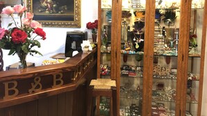 A wooden reception desk with a sign reading "B&B". Behind the desk, there is a framed painting, a coffee machine, and a vase with roses. To the right, there is a glass display case filled with various items. - Dream (Mestre)