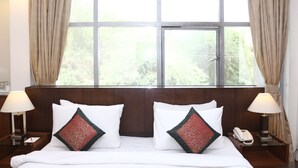 Ein Hotelzimmer mit großem Fenster, zwei Nachttischen mit Lampen, einem Bett mit weißer Bettwäsche und Dekokissen sowie einem Telefon auf dem rechten Tisch.