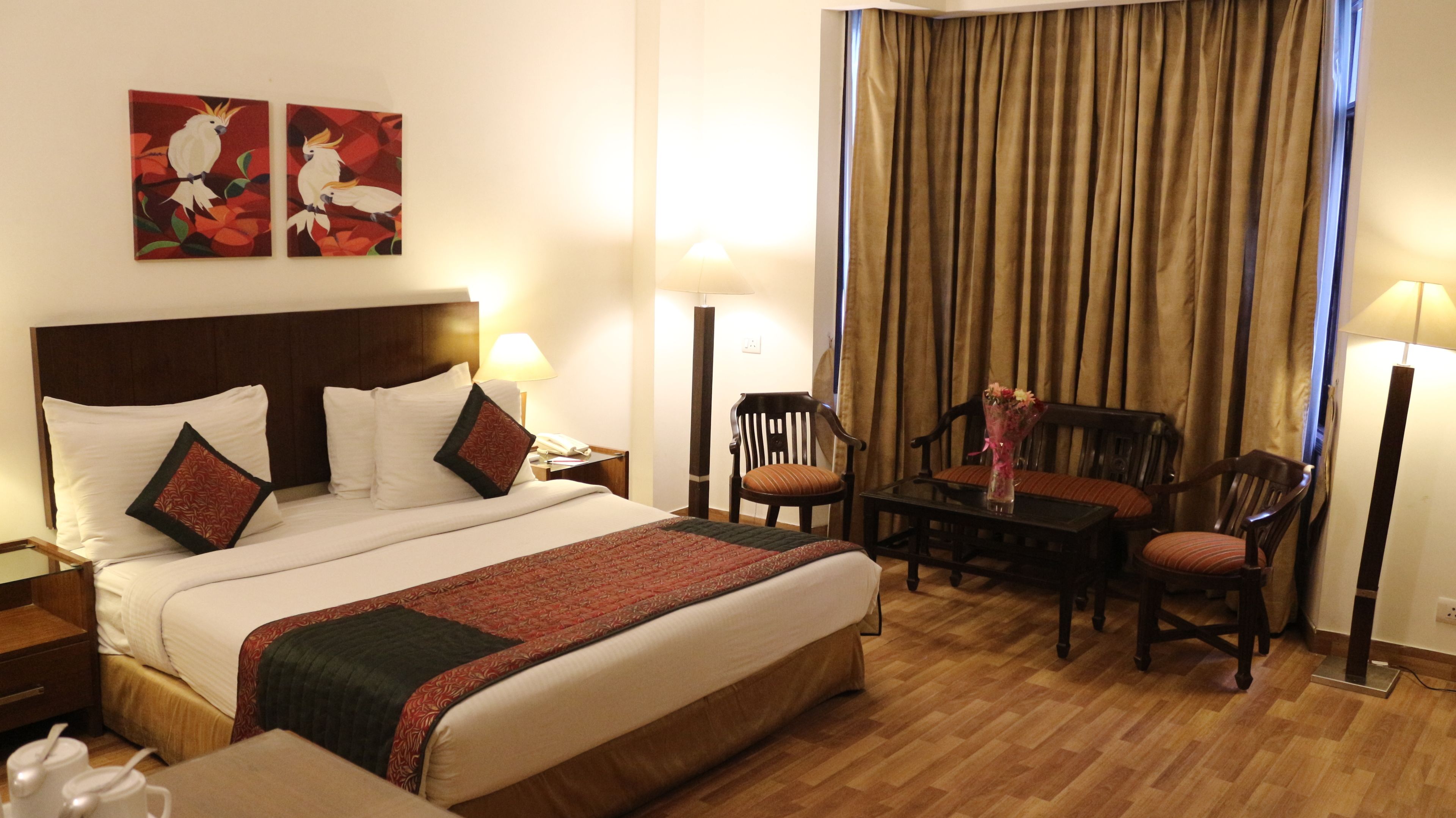 Habitación de hotel con una cama grande, dos cuadros, un escritorio con una silla, una mesa con un florero y una ventana con cortinas.