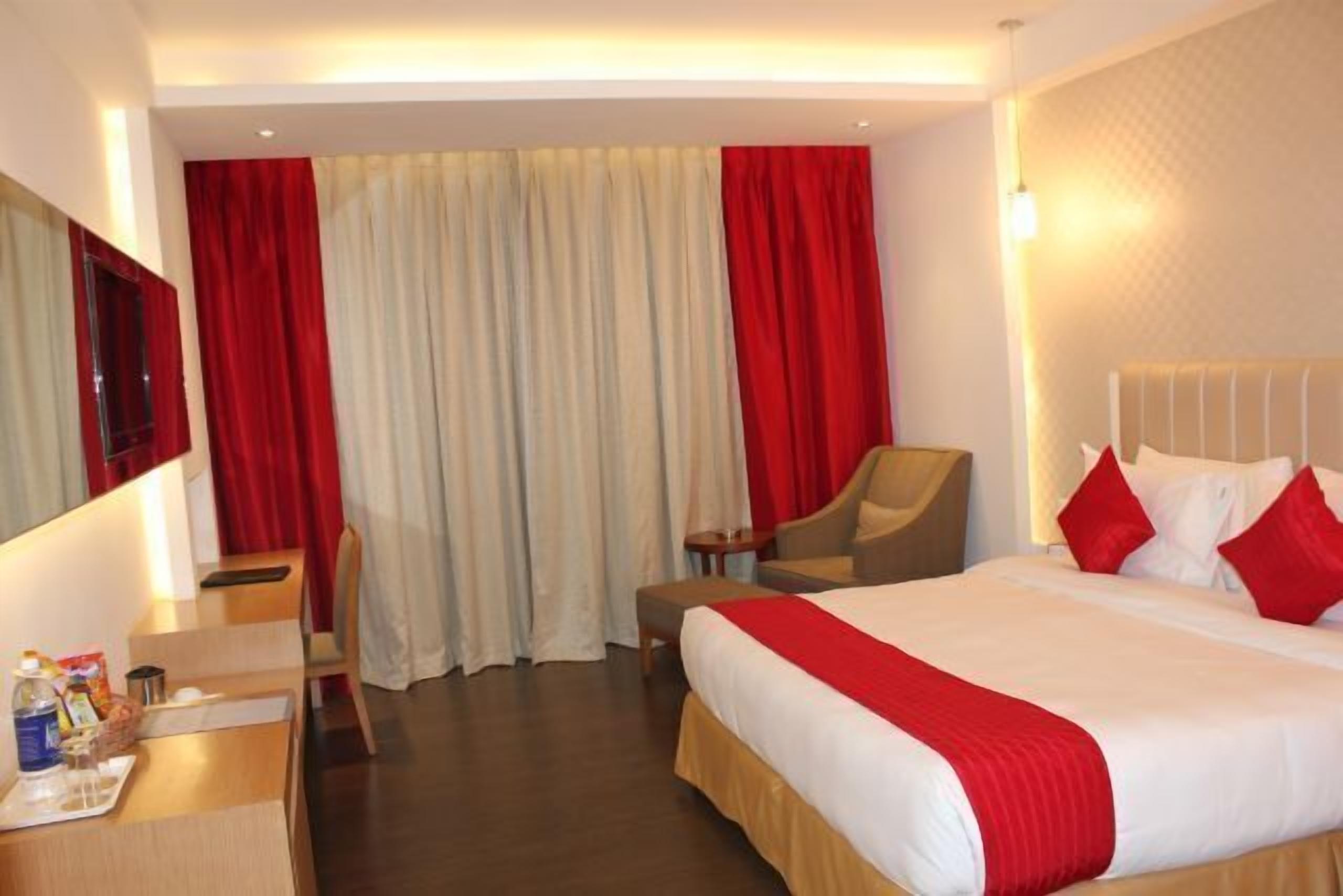 Ein Hotelzimmer mit einem großen Bett, einem Schreibtisch mit Stuhl, einem Fernseher und einem Fenster mit roten Vorhängen.