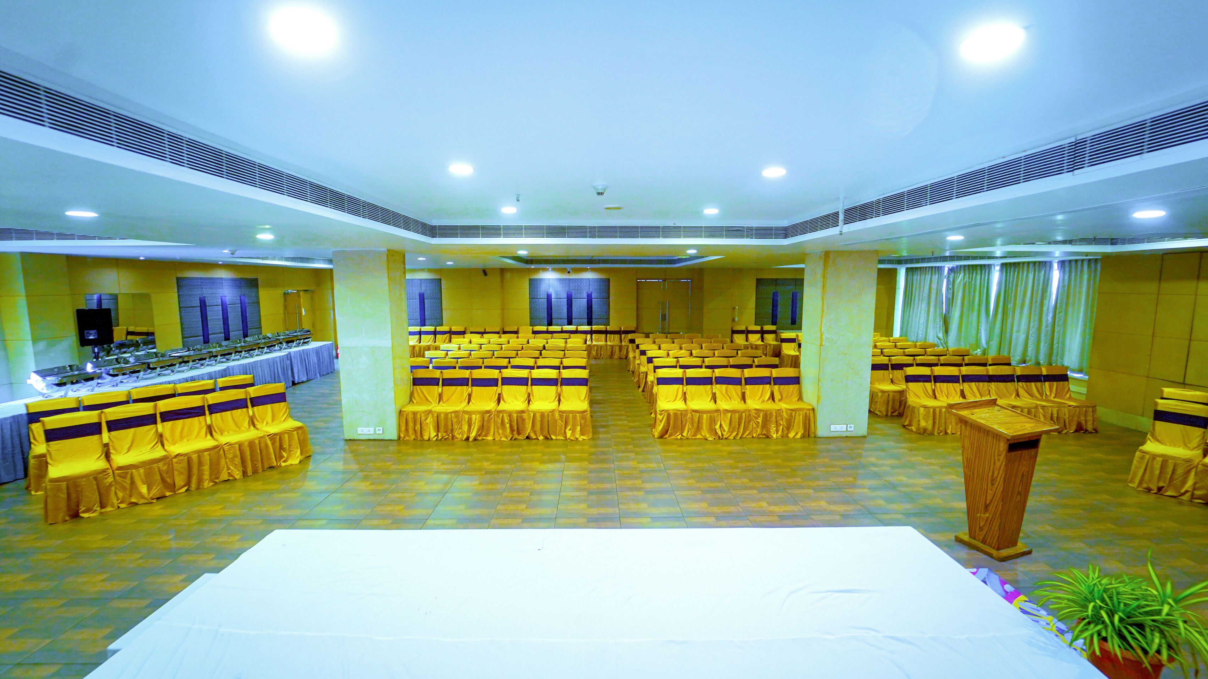 A large, well-lit banquet hall with rows of yellow chairs and tables, a stage at the front, and a long buffet table on the left.